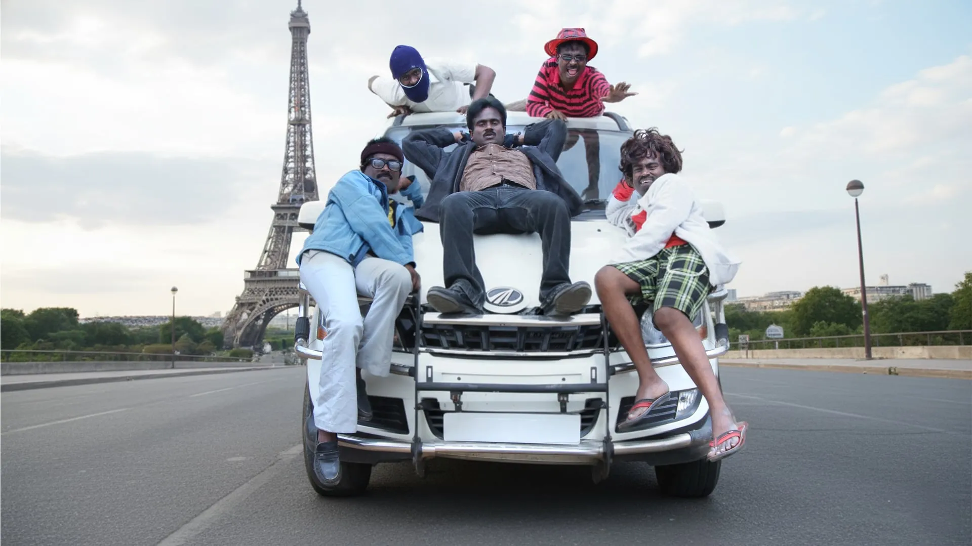 Sankagiri Rajkumar as five different characters lounging on a white SUV parked in front of the Eiffel Tower