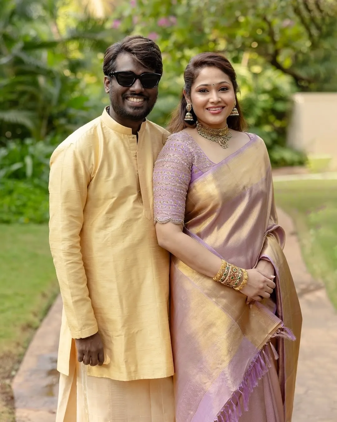 Filmmaker Atlee in a yellow silk kurta and Priya Atlee in a lavender and gold kanjivaram saree, standing close together for a posed garden portrait
