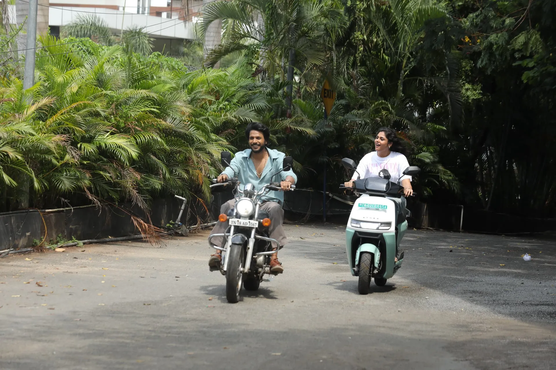A man on a motorbike and a woman on a scooter ride side by side down a leafy driveway in Sigma