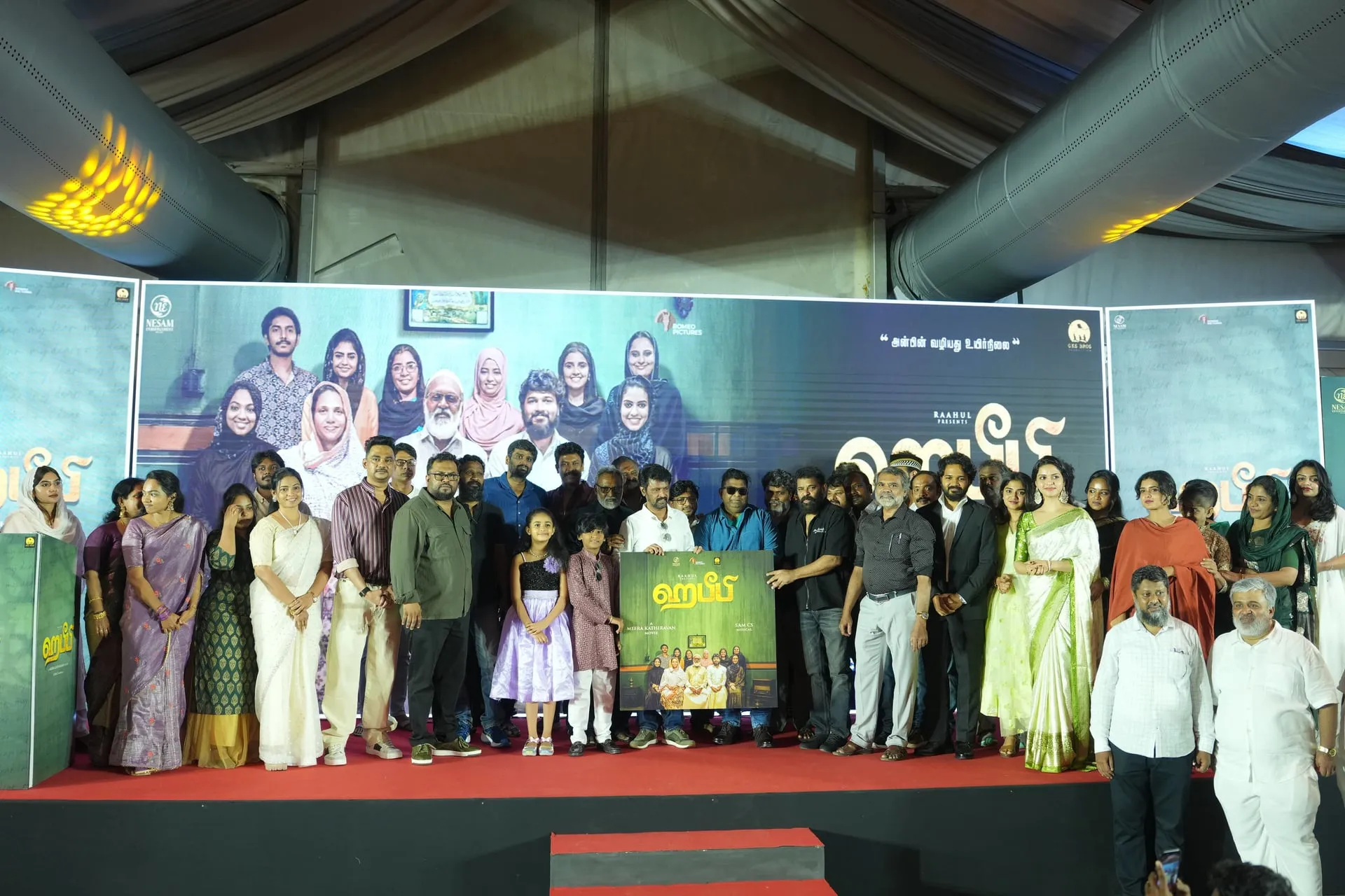 Wide stage photograph at the Habeebi music launch, with the full cast and crew of the film and a row of Tamil filmmakers including Meera Kathiravan, Kasturi Raja, Malavika Manoj and several directors lined up around the unveiled audio album poster