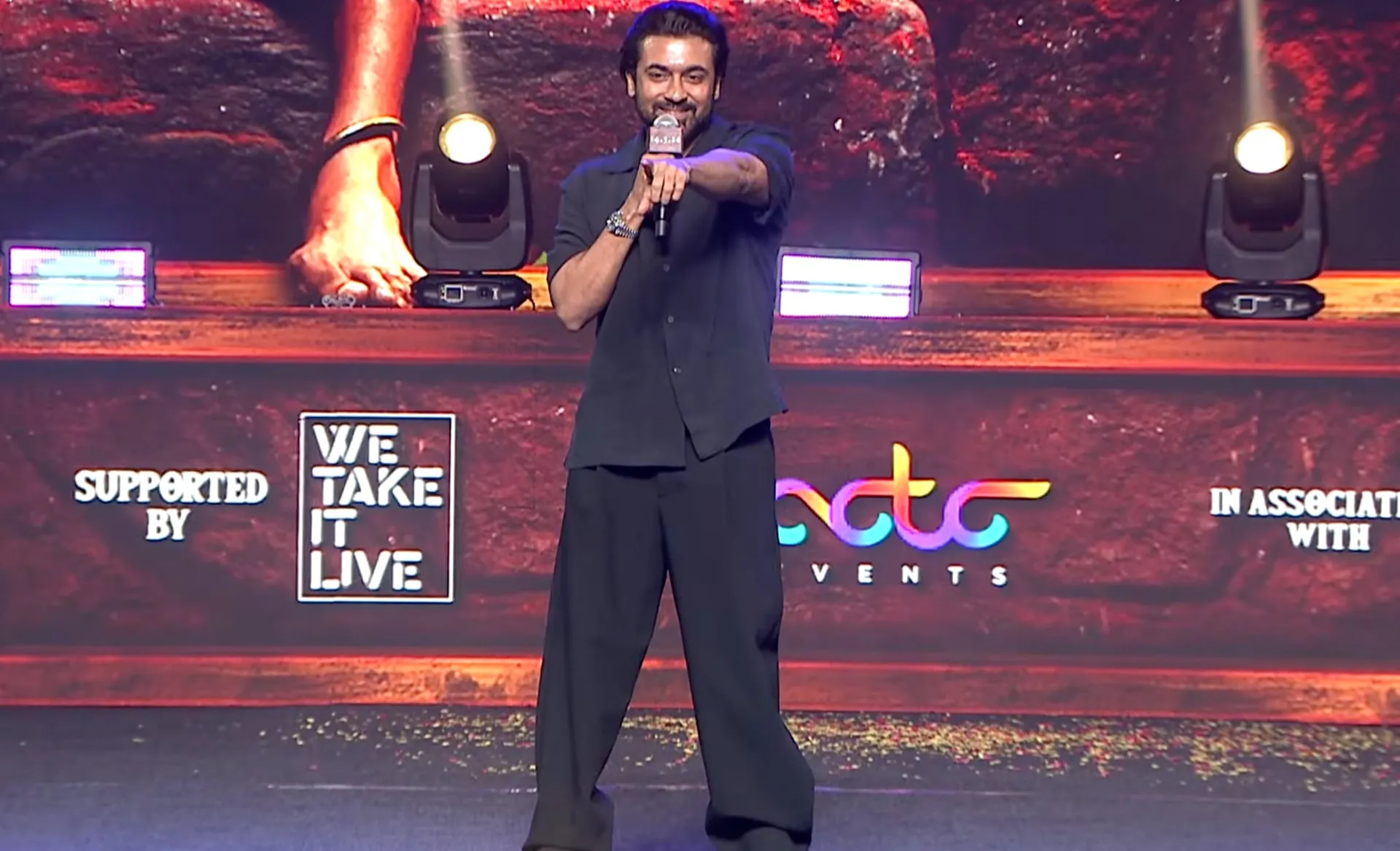 Suriya in a plain black shirt and trousers on stage at the Karuppu pre-release event in Hyderabad, mid-speech with mic in hand