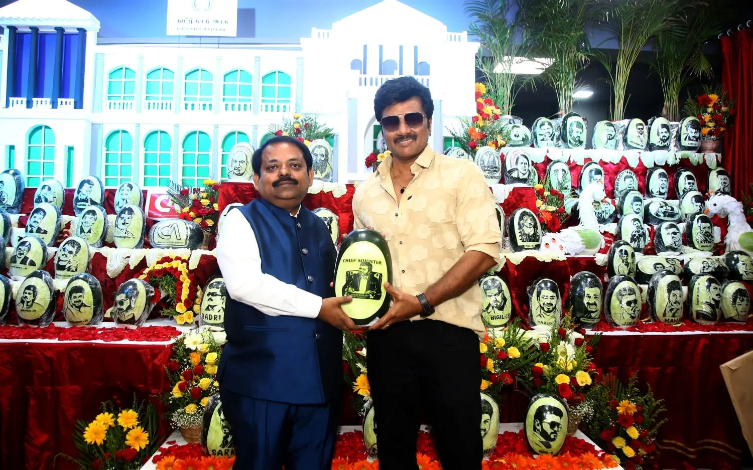 Two men hold a carved ash gourd labelled Chief Minister in front of a tiered display of Vijay portraits