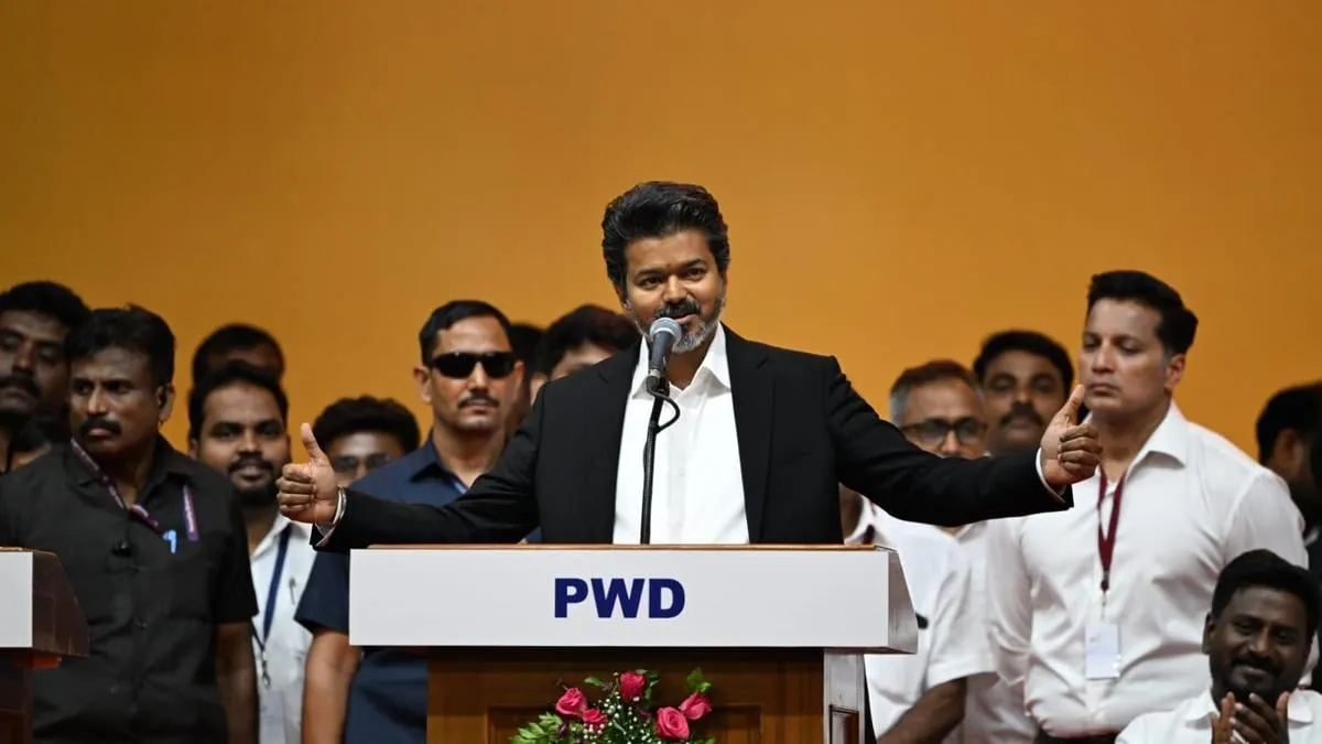 Vijay at a podium with PWD signage, microphone in hand, raising both thumbs as security and supporters stand behind him