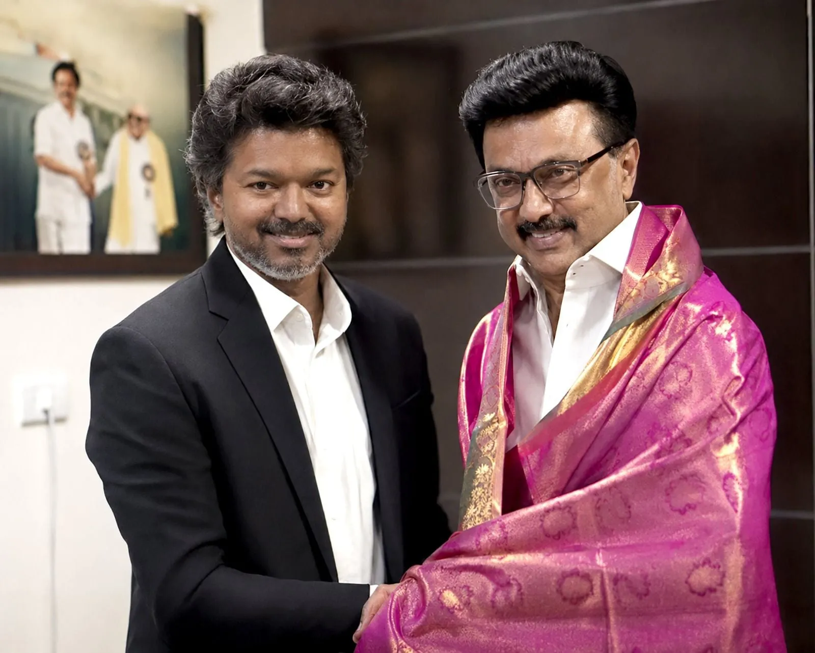 TN Chief Minister Vijay shakes hands with predecessor MK Stalin at Stalin's Chennai residence, with Stalin draped in a pink-and-gold silk shawl and a Karunanidhi portrait on the wall behind