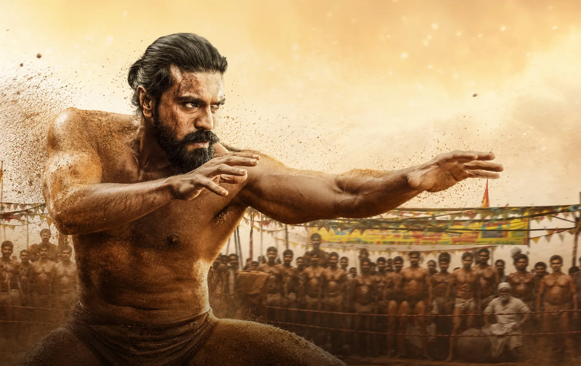 Ram Charan, mud-caked and shirtless, crouches in a wrestling stance inside a packed akhada in Peddi