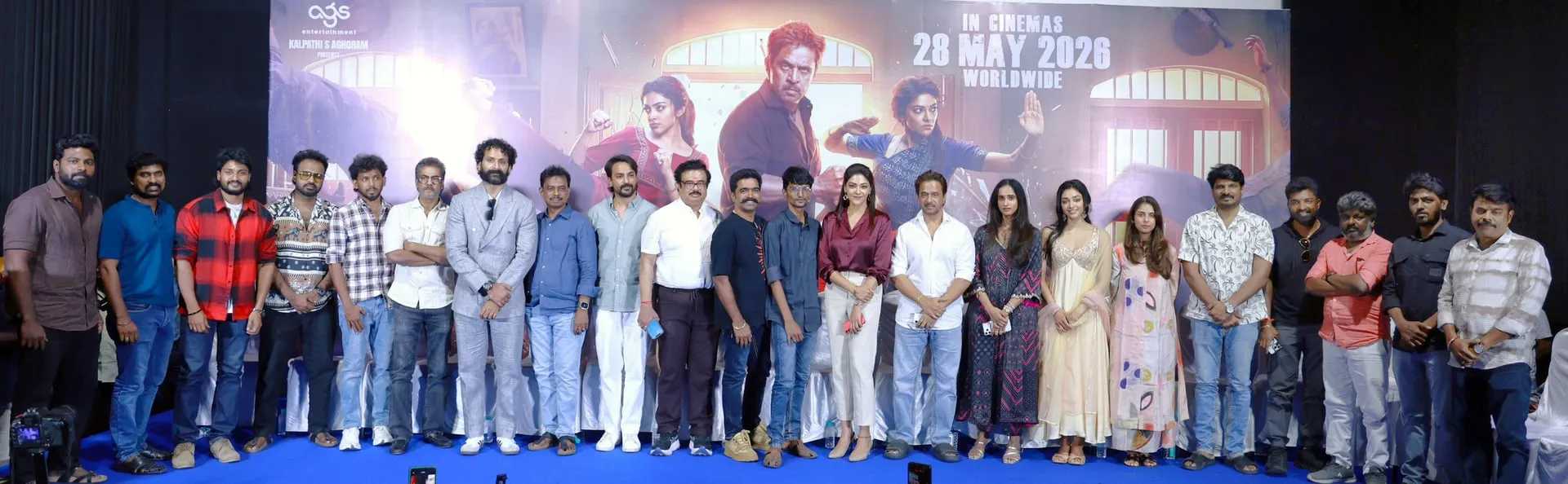 Wide stage photograph at the Blast pre-release event, with Arjun, Abhirami, Preithy Mukundhan, debut director Subash K Raj, composer Ravi Basrur and the rest of the cast and crew lined up in front of the Blast key art and the AGS Entertainment logo, with 'In Cinemas 28 May 2026 Worldwide' marked across the backdrop