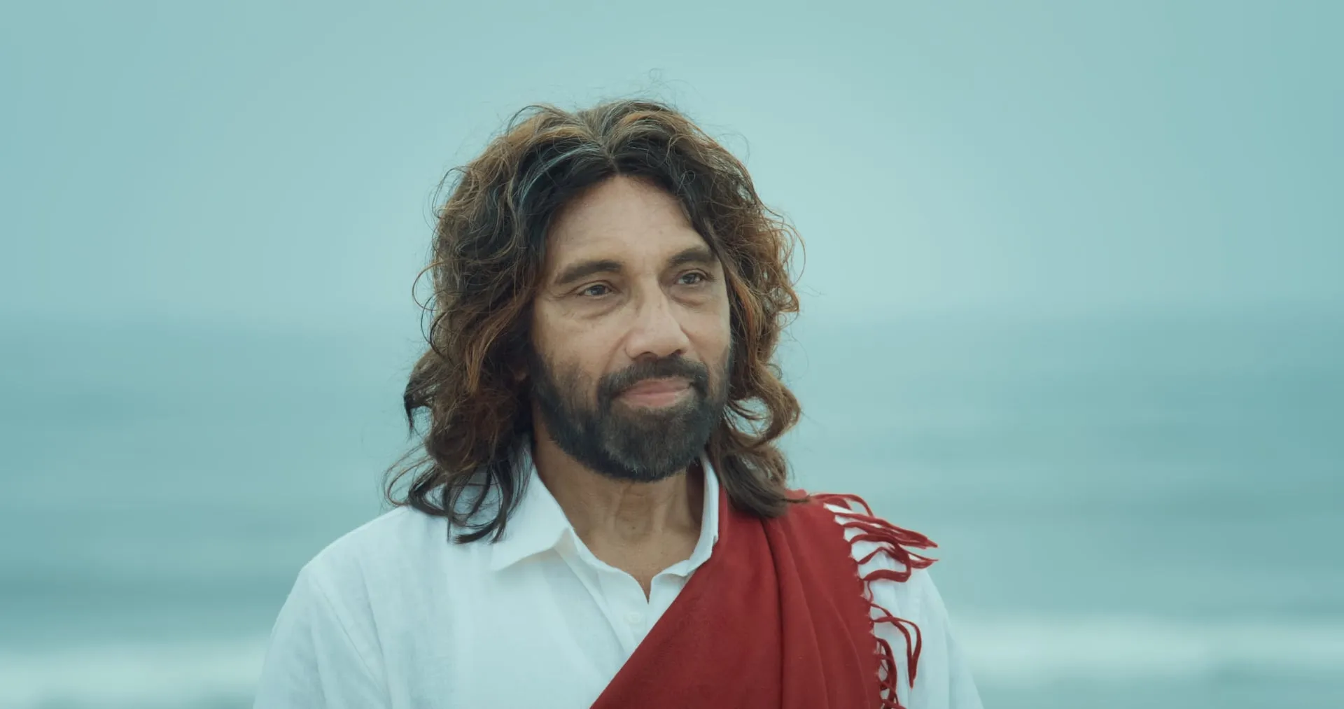 A long-haired man in a white shirt and red shawl stands on a misty beach, looking calmly toward the camera.