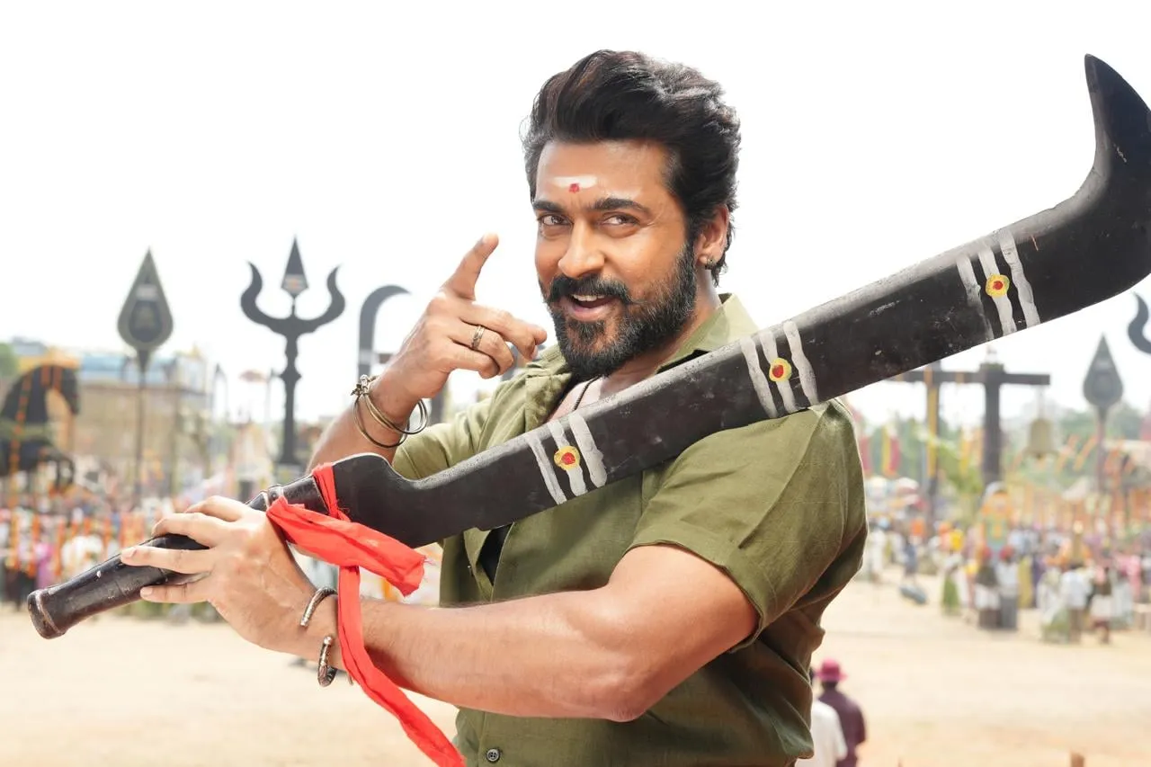 Suriya in Karuppu, in an olive shirt holding a temple-painted aruval, grinning at the camera with a Karuppasamy procession in the deep background