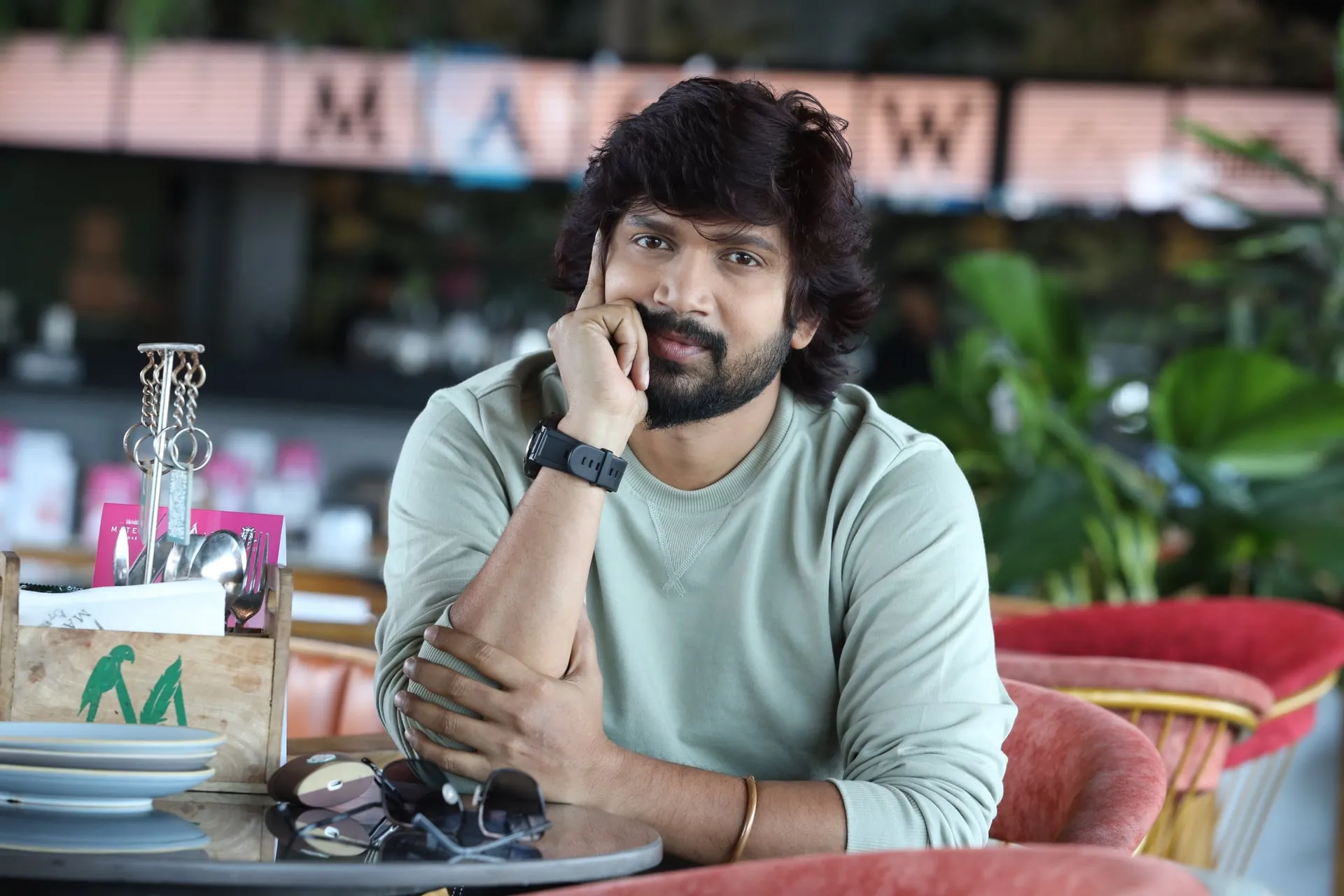 Vijay Vishwa seated at a cafe table in a sage sweatshirt, chin resting on his hand, looking into the camera
