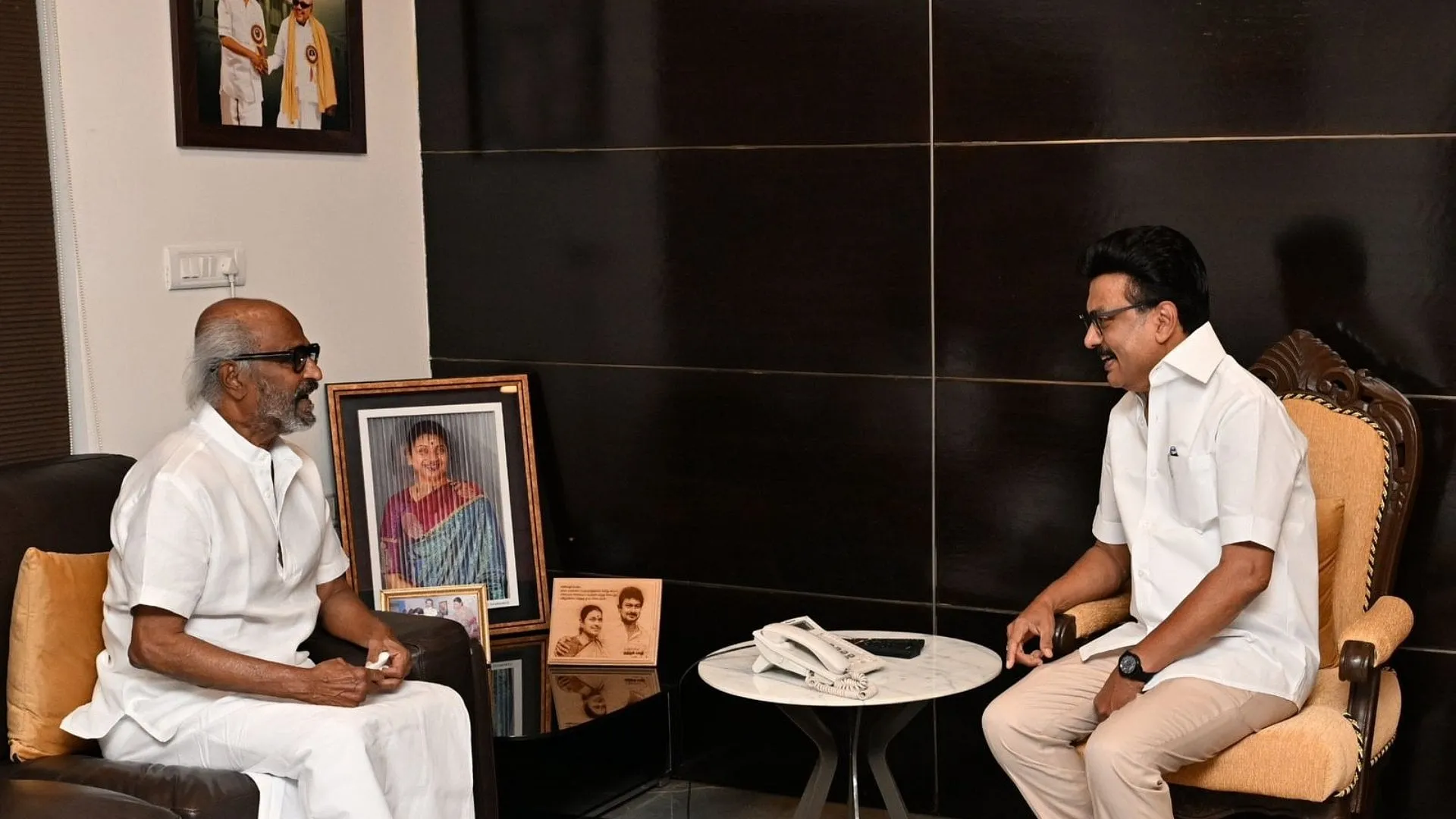 Rajinikanth in white seated opposite MK Stalin in his residence in a quiet face-to-face conversation, framed by family photographs
