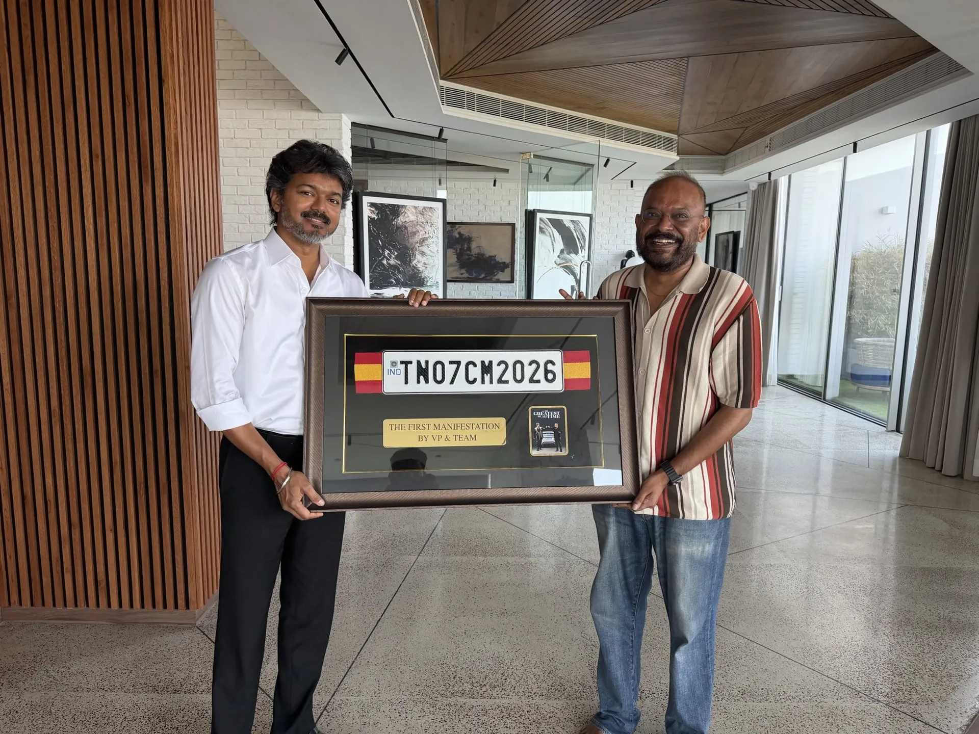 Chief Minister Vijay in a white shirt and dark trousers and director Venkat Prabhu in a brown-striped polo and jeans, both standing in the CM's residence and holding a large brown-framed display that contains the original TN07CM2026 number plate from GOAT, a small brass plaque reading THE FIRST MANIFESTATION BY VP AND TEAM, and a miniature GOAT poster