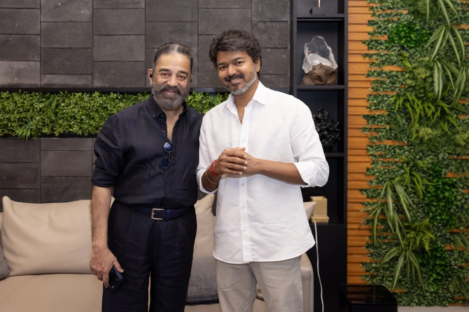 Kamal Haasan in navy linen with the chief minister Vijay in white at Vijay's Neelangarai residence, standing side by side and smiling, with a textured slate wall and a green vertical garden behind them