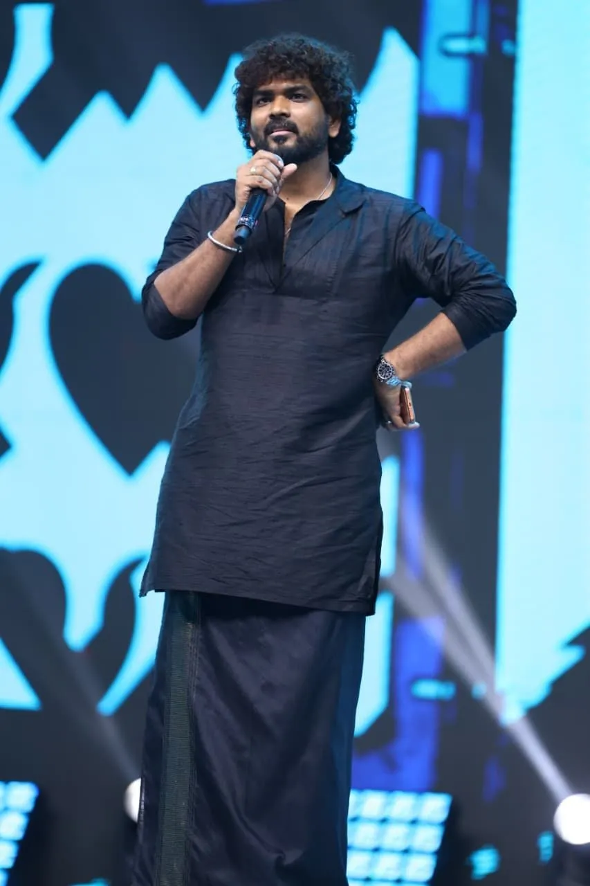 Vignesh Shivan in a black kurta and veshti, on stage at the Love Insurance Kompany pre-release event, holding a mic