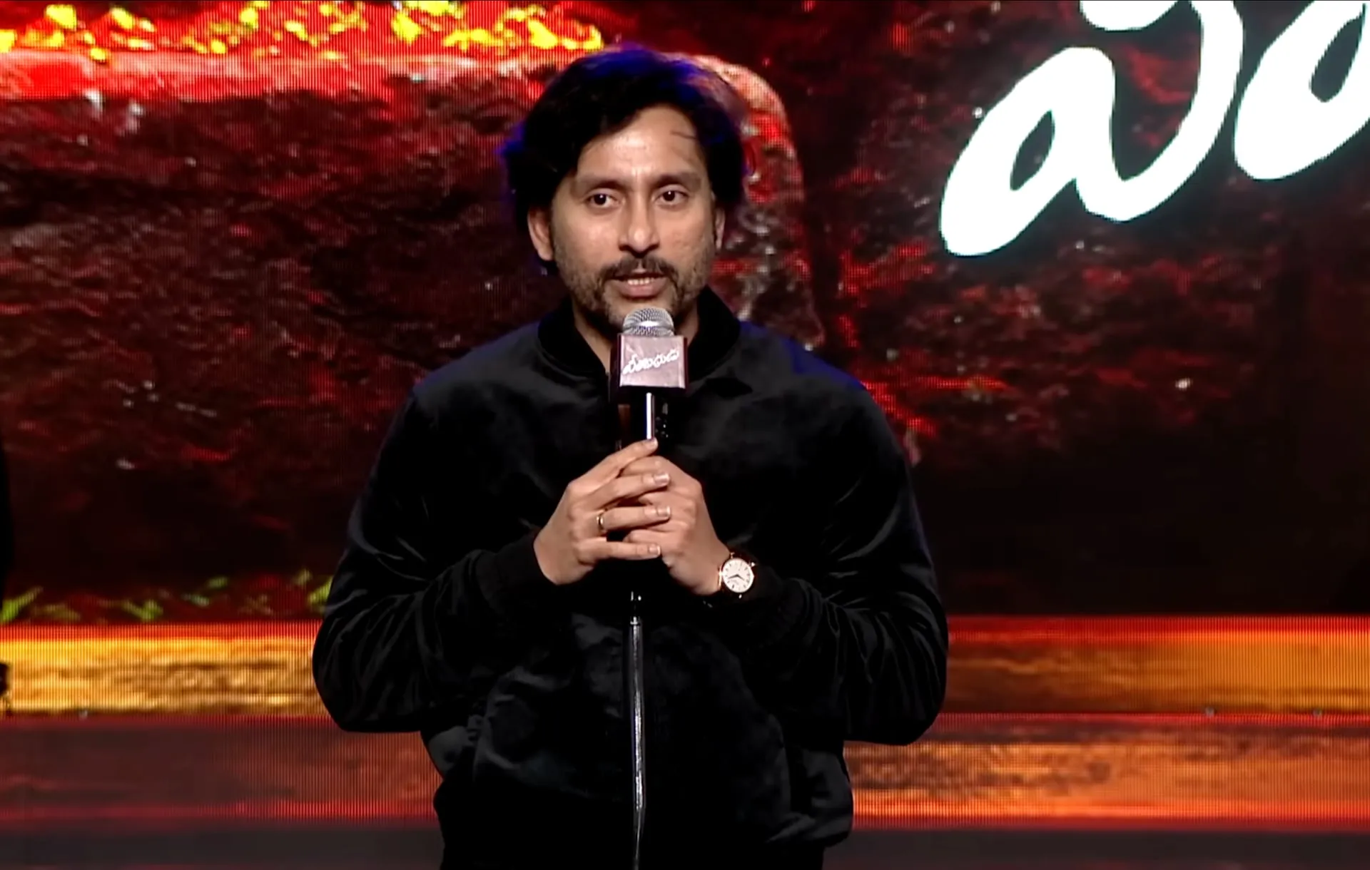 RJ Balaji in a black velvet jacket on stage at the Veerabadhrudu pre-release event in Hyderabad, speaking into a mic in front of a red backdrop