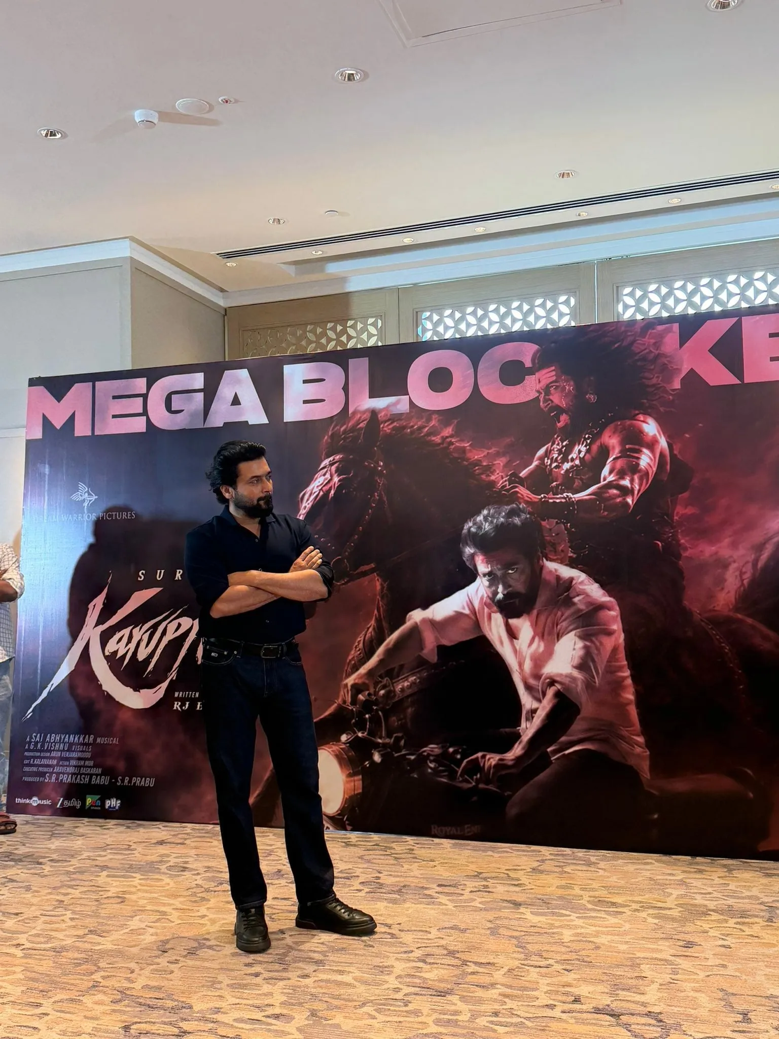 RJ Balaji standing in front of a Karuppu blockbuster banner featuring Suriya