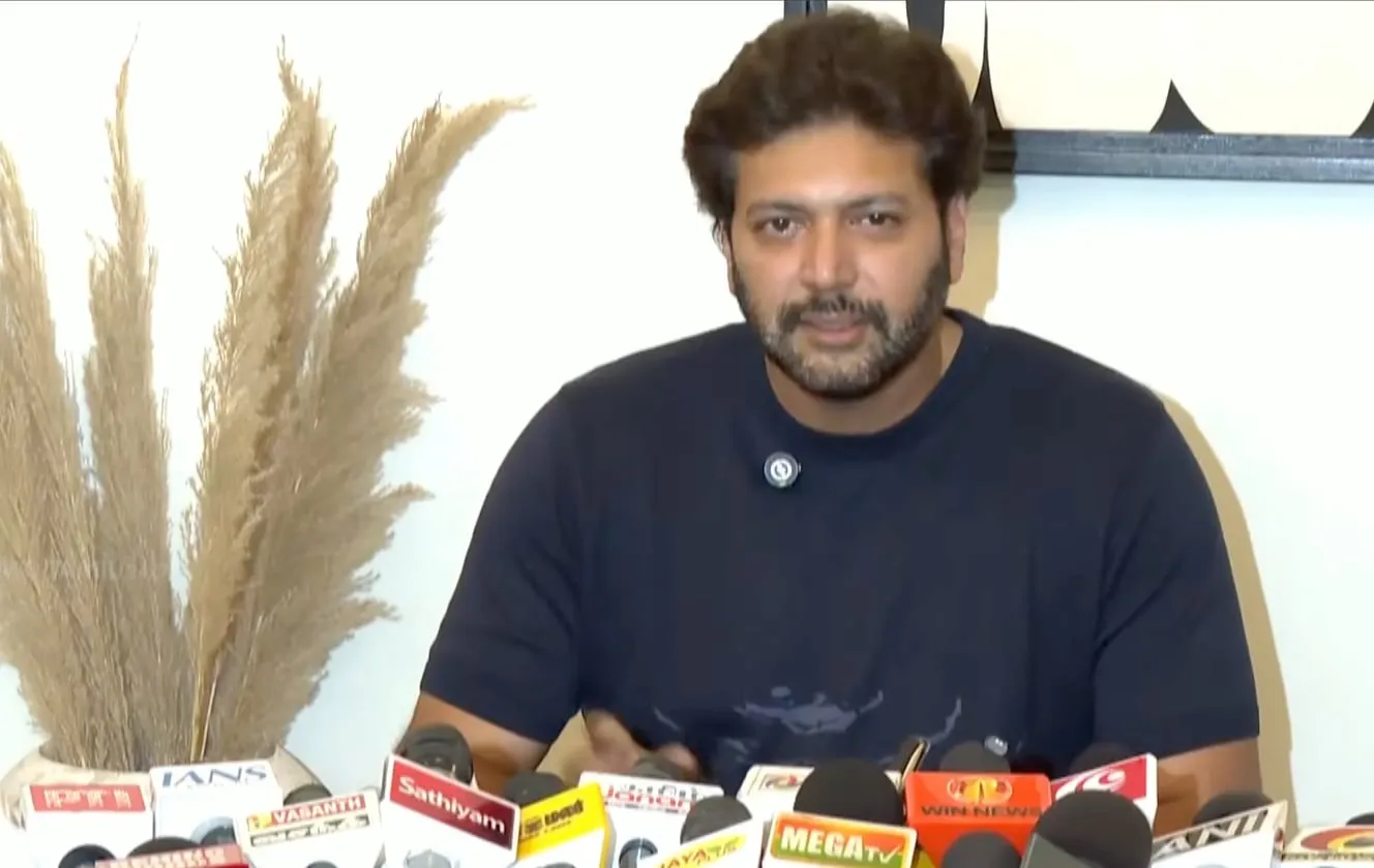 Ravi Mohan in a navy T-shirt seated behind a bank of channel microphones at his office, addressing reporters about his ongoing divorce