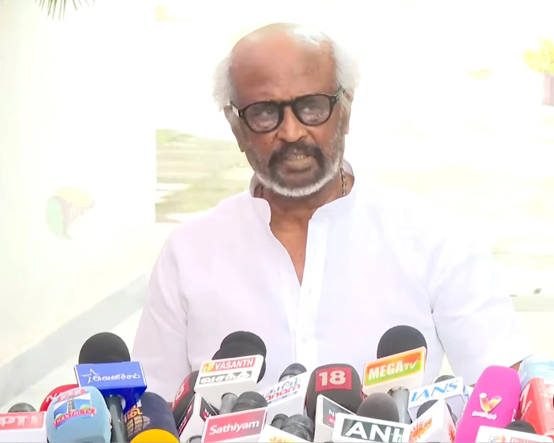 Rajinikanth in a white kurta and dark glasses standing behind a bank of channel microphones at his Poes Garden residence, addressing reporters about the new chief minister Vijay