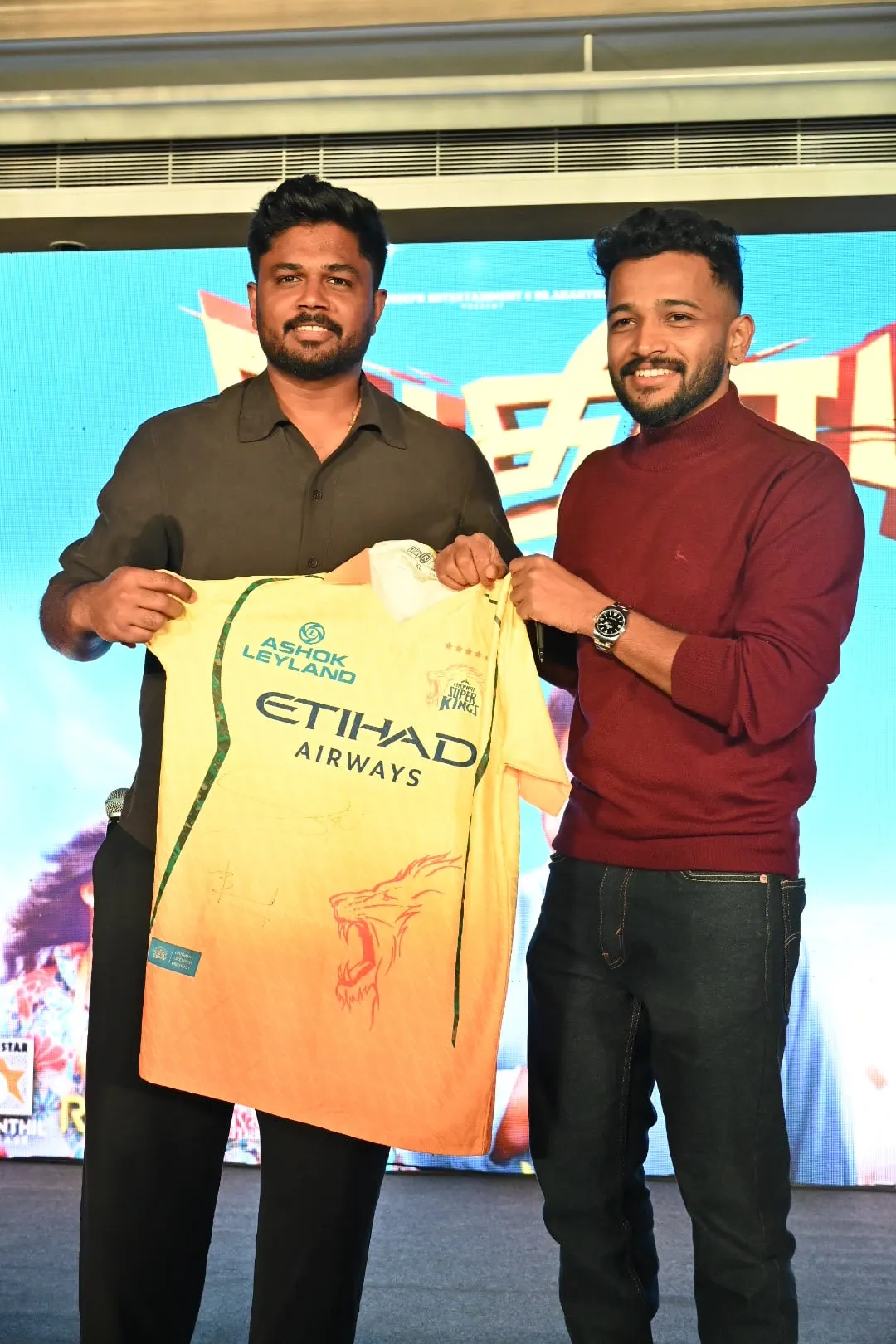 Sanju Samson in a grey shirt standing on the left, Basil Joseph in a maroon sweater on the right holding up a yellow Chennai Super Kings jersey he has just signed, both posed against the blue Athiradi backdrop at the Chennai pre-release event