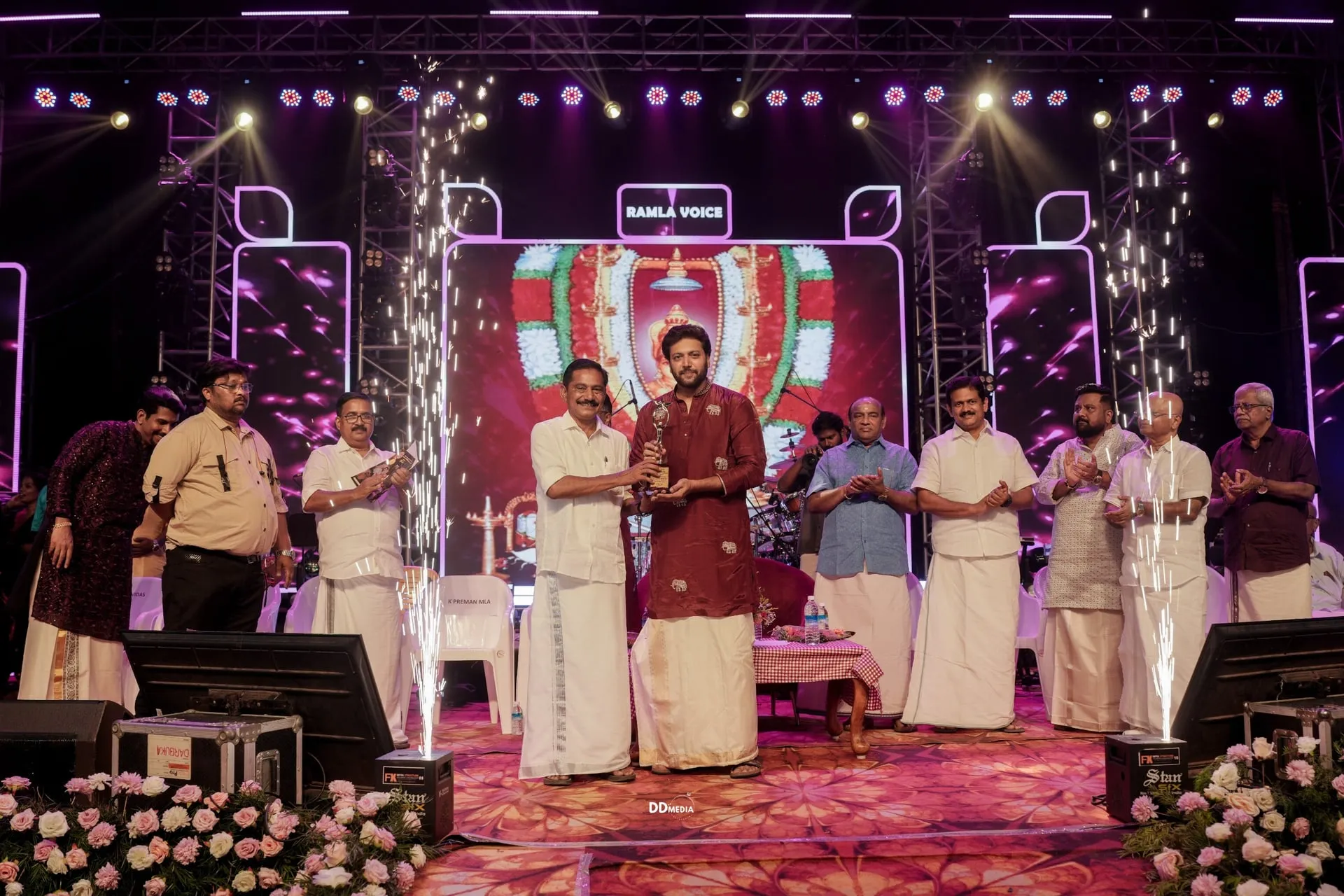 Ravi Mohan in a maroon kurta receiving the Devi Ratna trophy on stage at the Pallassana Pazhayakavu Sree Meenkulathi Bhagavathi Temple in Palakkad, flanked by trustees and dignitaries, with sparklers in the foreground