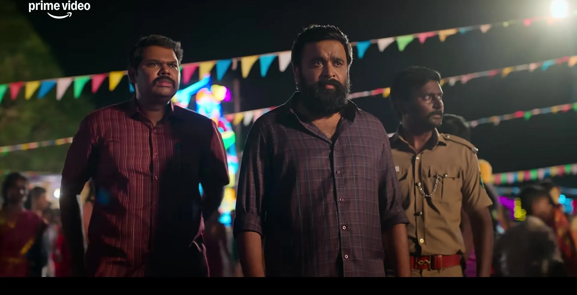 M Sasikumar as SI Moosa Raaza walks through a village festival at night flanked by two colleagues, with strings of colourful flags overhead