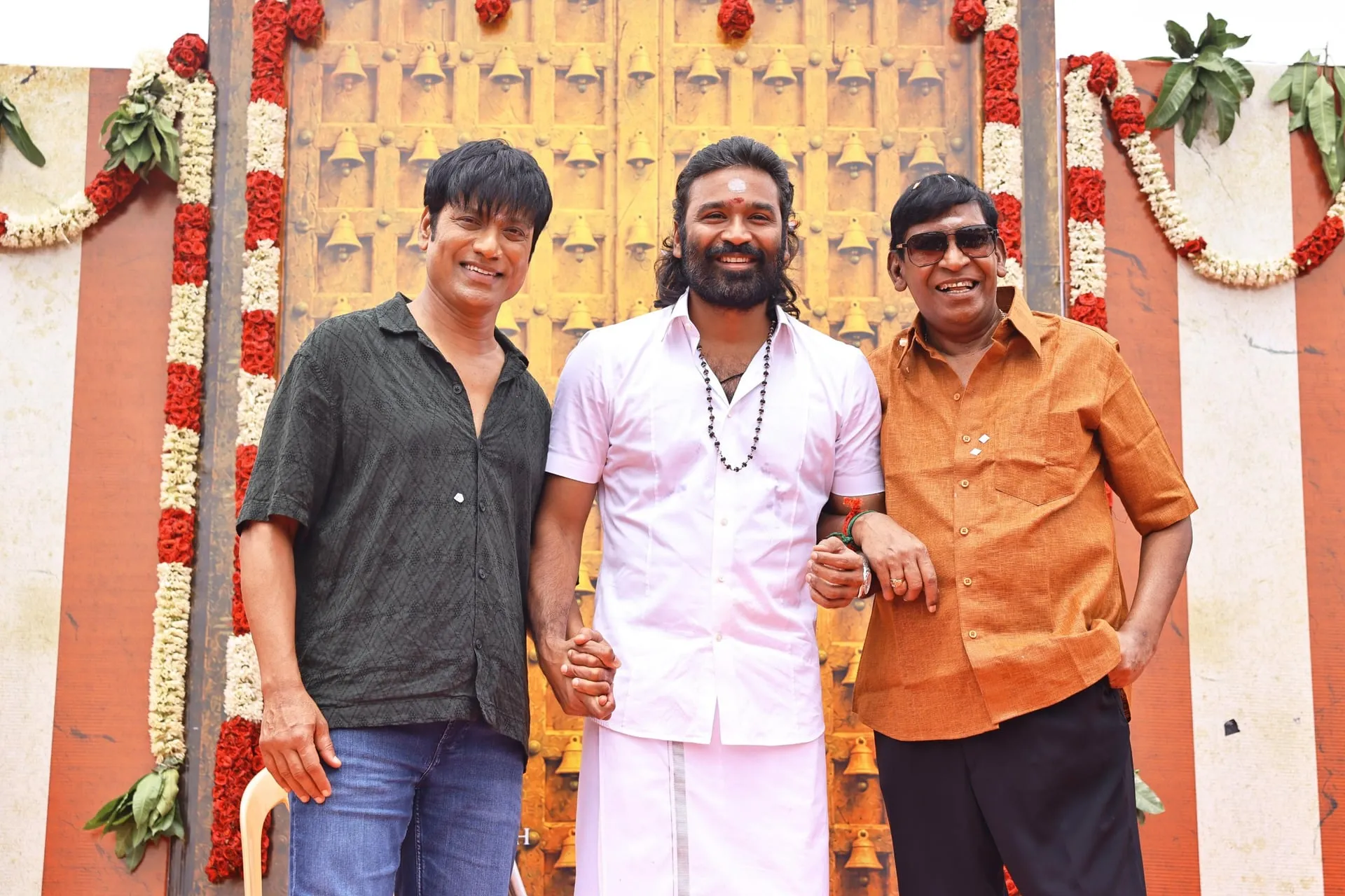 Dhanush in a white shirt and veshti with members of the D56 team in front of a flower-decked temple door at the film's pooja