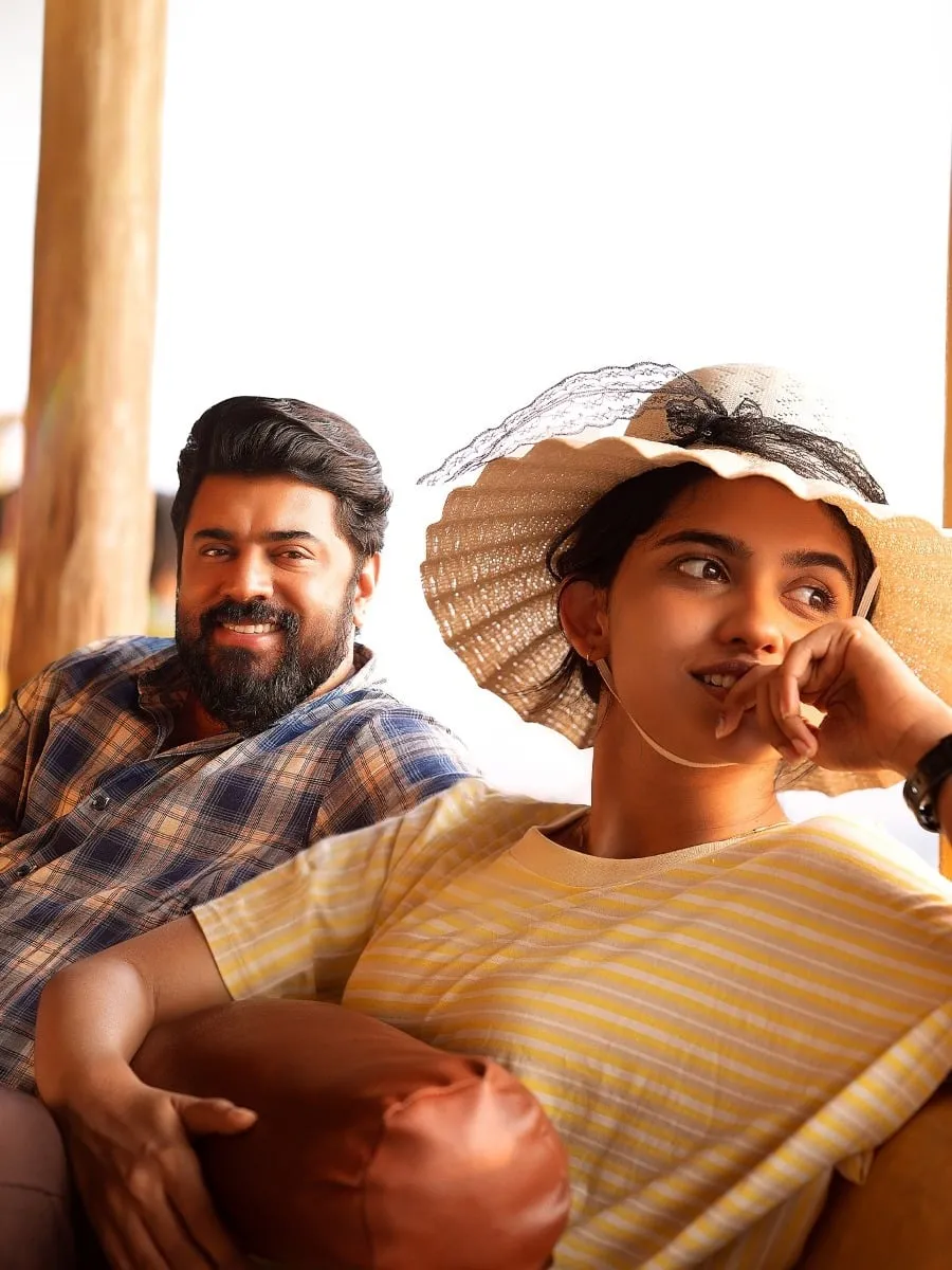 Nivin Pauly in a plaid shirt grins beside Mamitha Baiju in a wide straw hat as they sit on a sunlit sofa in Bethlehem Kudumba Unit