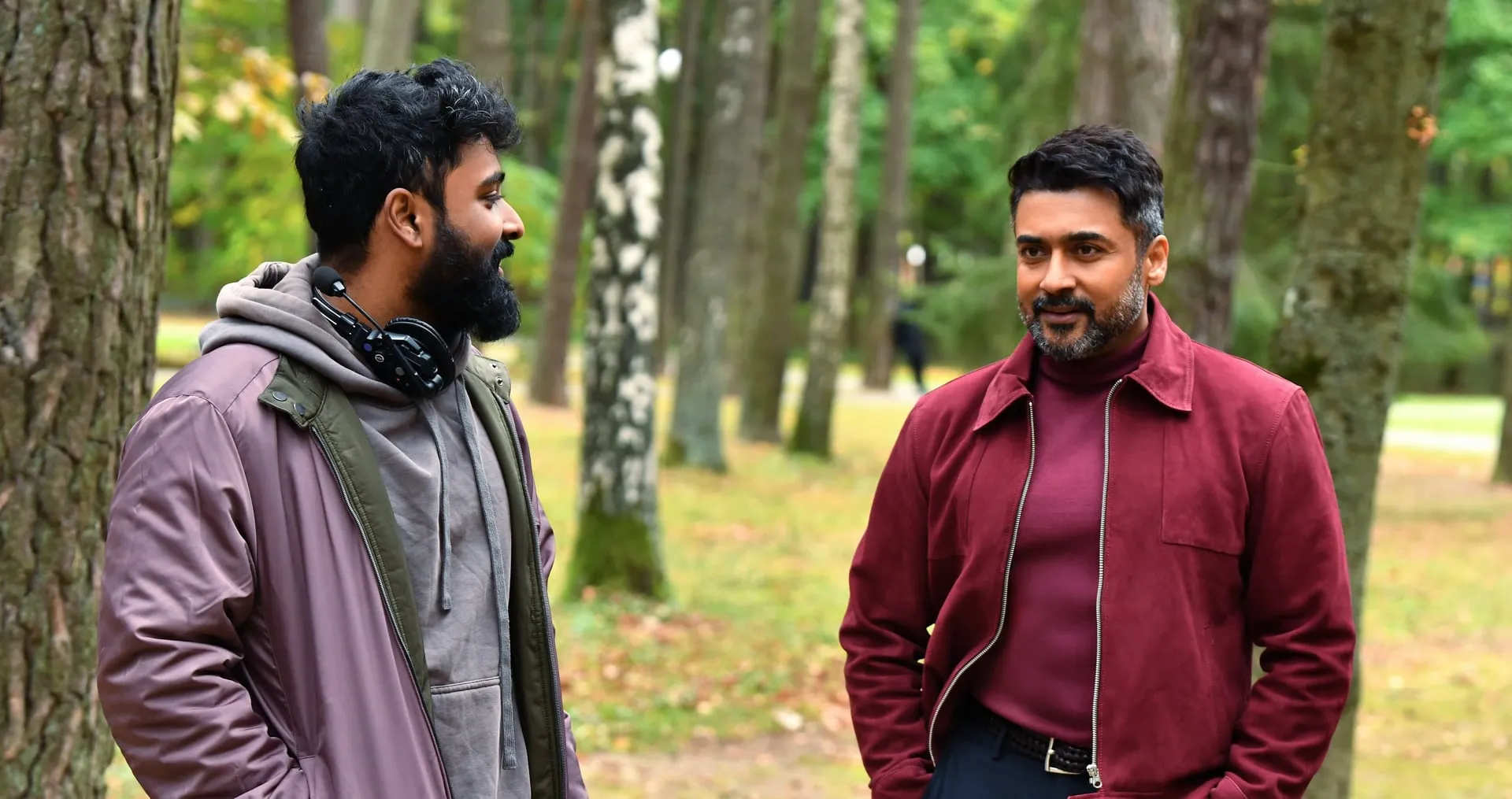 Suriya in a maroon suede jacket chats with a crew member in a birch grove on the set of Vishwanath & Sons