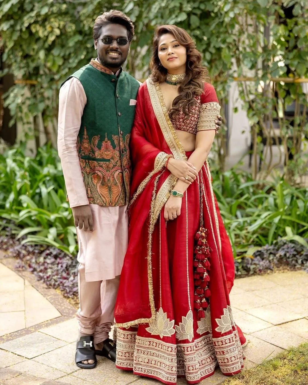 Atlee in a green sleeveless Nehru jacket over a cream kurta, with Priya in a deep red lehenga set, standing for a posed garden portrait at a family function