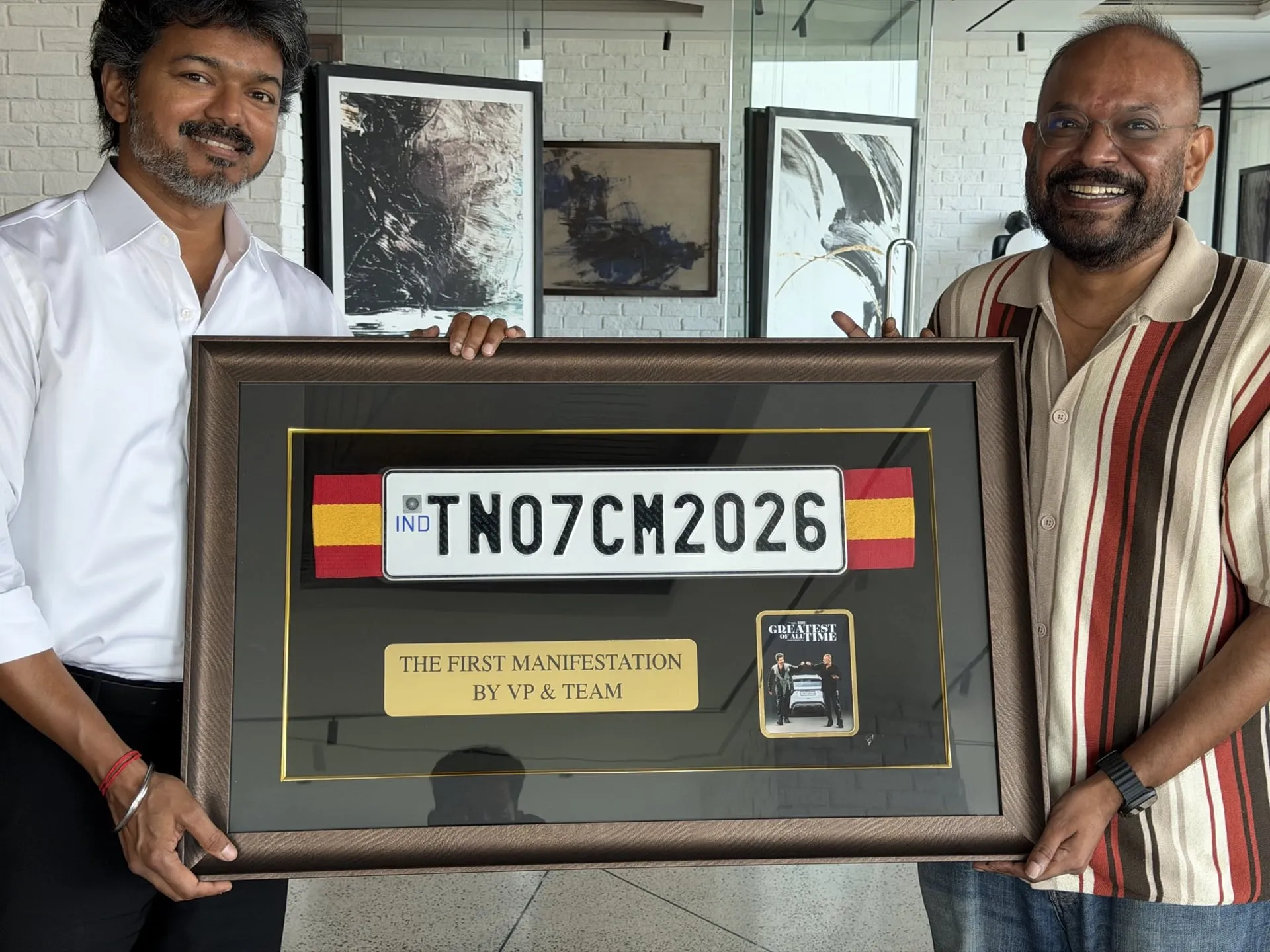 A close-up of the framed gift, with the IND-marked TN07CM2026 number plate set above a brass plaque reading THE FIRST MANIFESTATION BY VP & TEAM and a miniature GOAT poster, held between CM Vijay on the left and Venkat Prabhu on the right