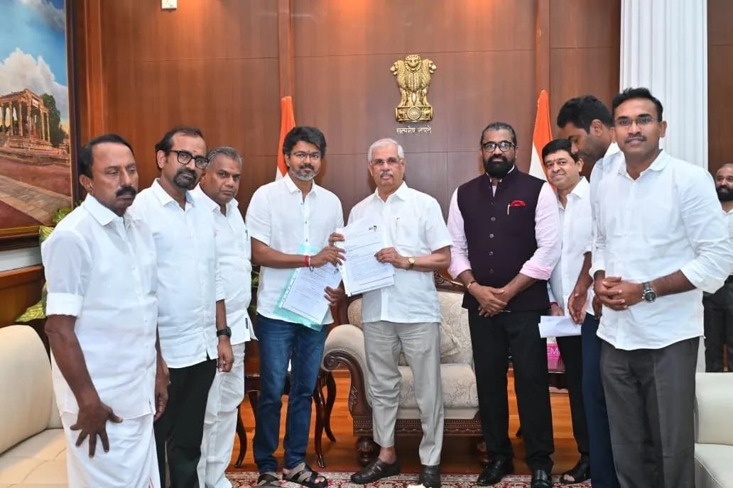 Vijay handing letters of support to Governor Arlekar at Lok Bhavan, both holding the document jointly, with the TVK delegation flanking them under the national emblem