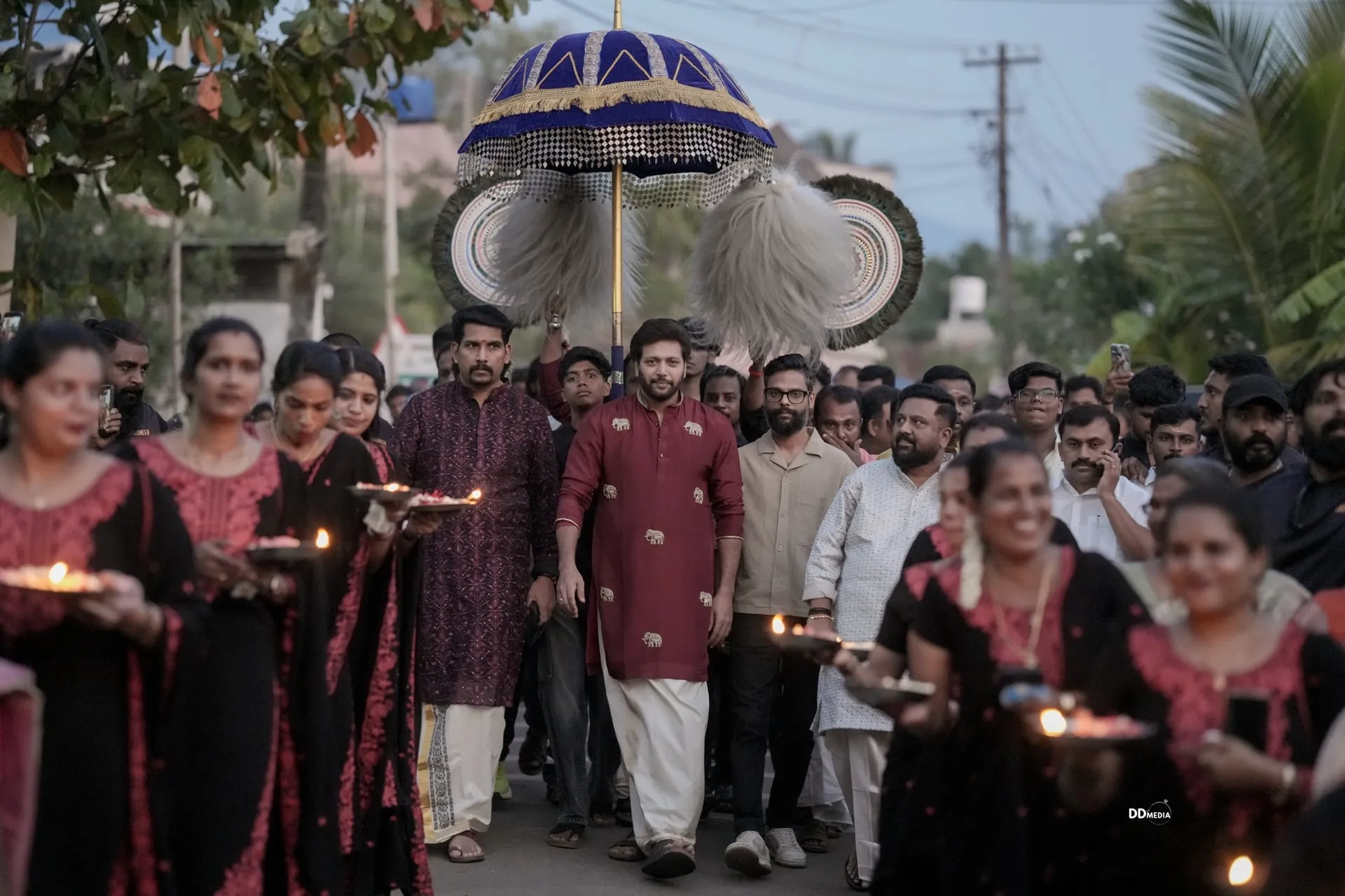 Ravi Mohan walks the customary temple procession into the Utsav 2026 grounds, in a maroon kurta under a ceremonial umbrella, with locals carrying lamps along the route.