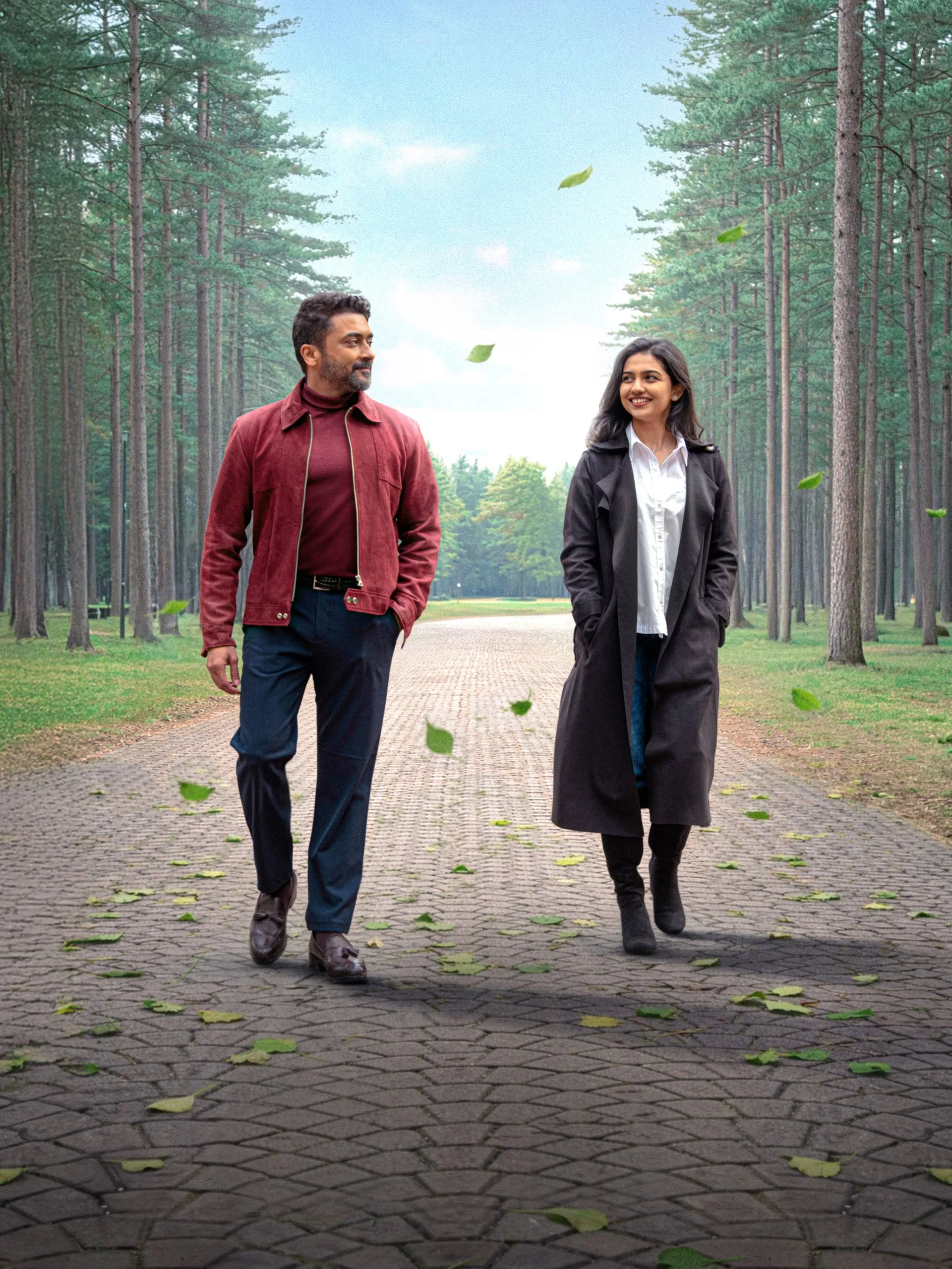 Suriya and Mamitha Baiju walking side by side down a leaf-strewn path lined with tall pines