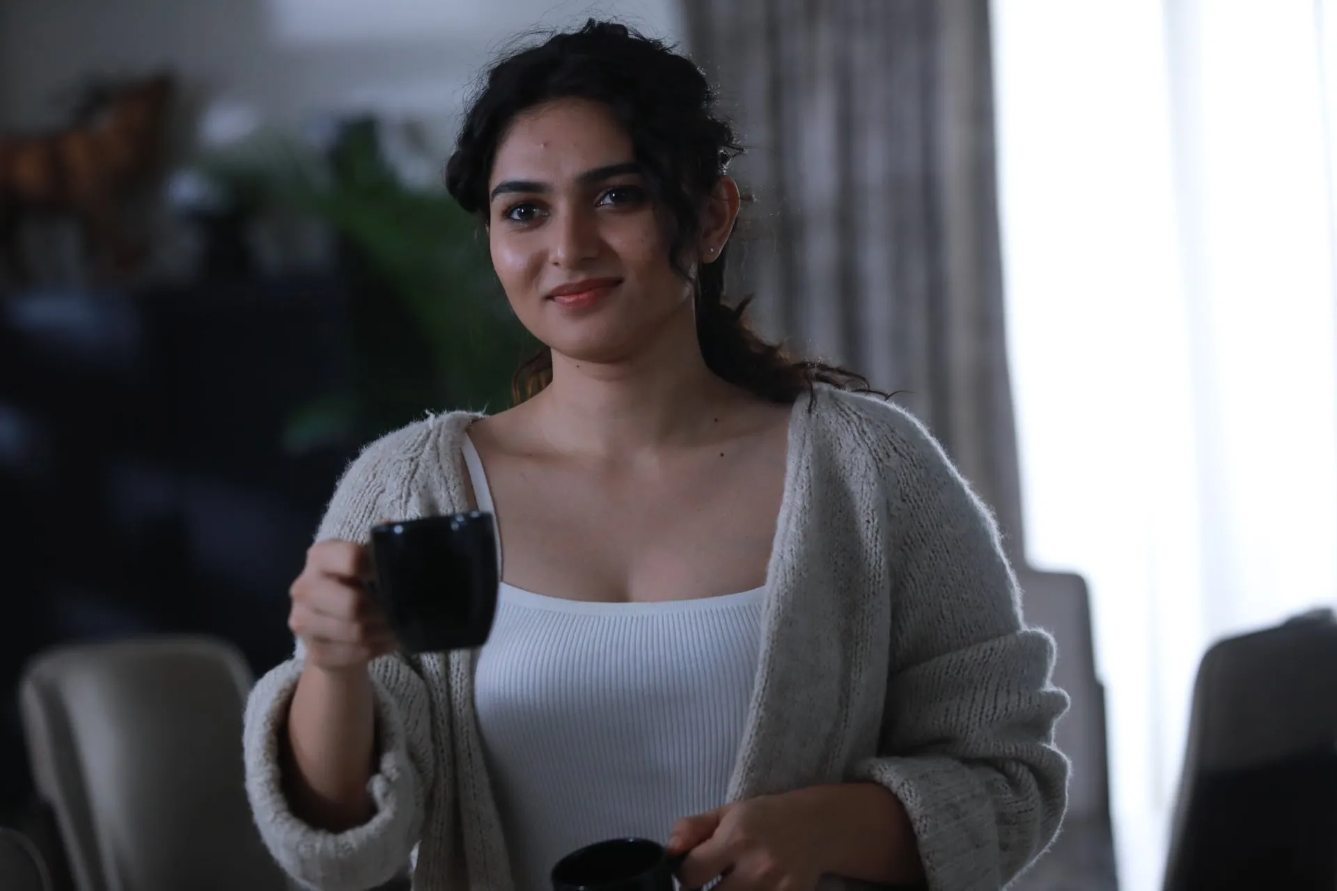 Kayadu Lohar as Sindhu in a cream cardigan holding a black coffee mug and smiling in the apartment in Immortal