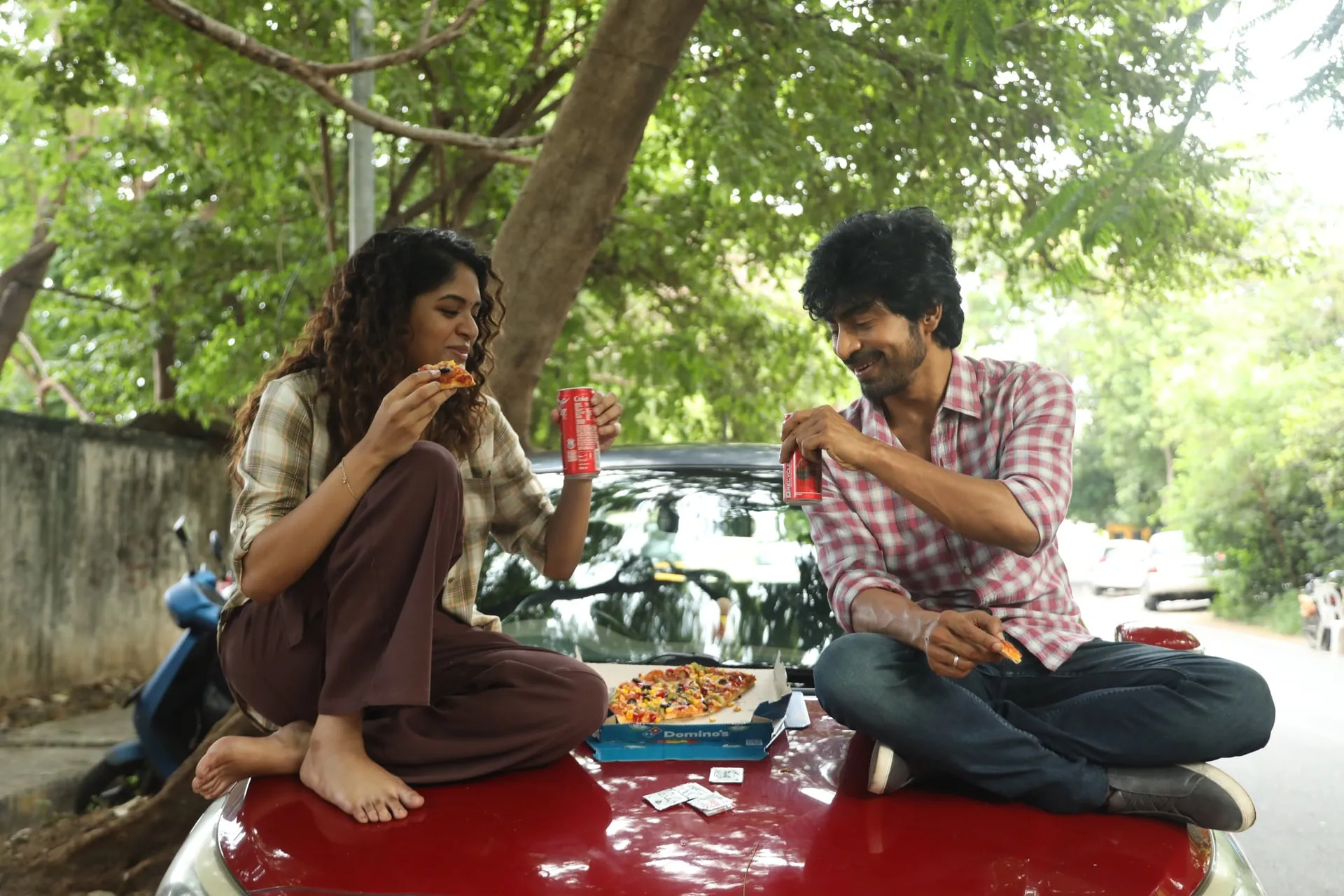 Anjali and Raghu sit cross-legged on a red car bonnet under a tree, sharing pizza and cold drinks in Once More