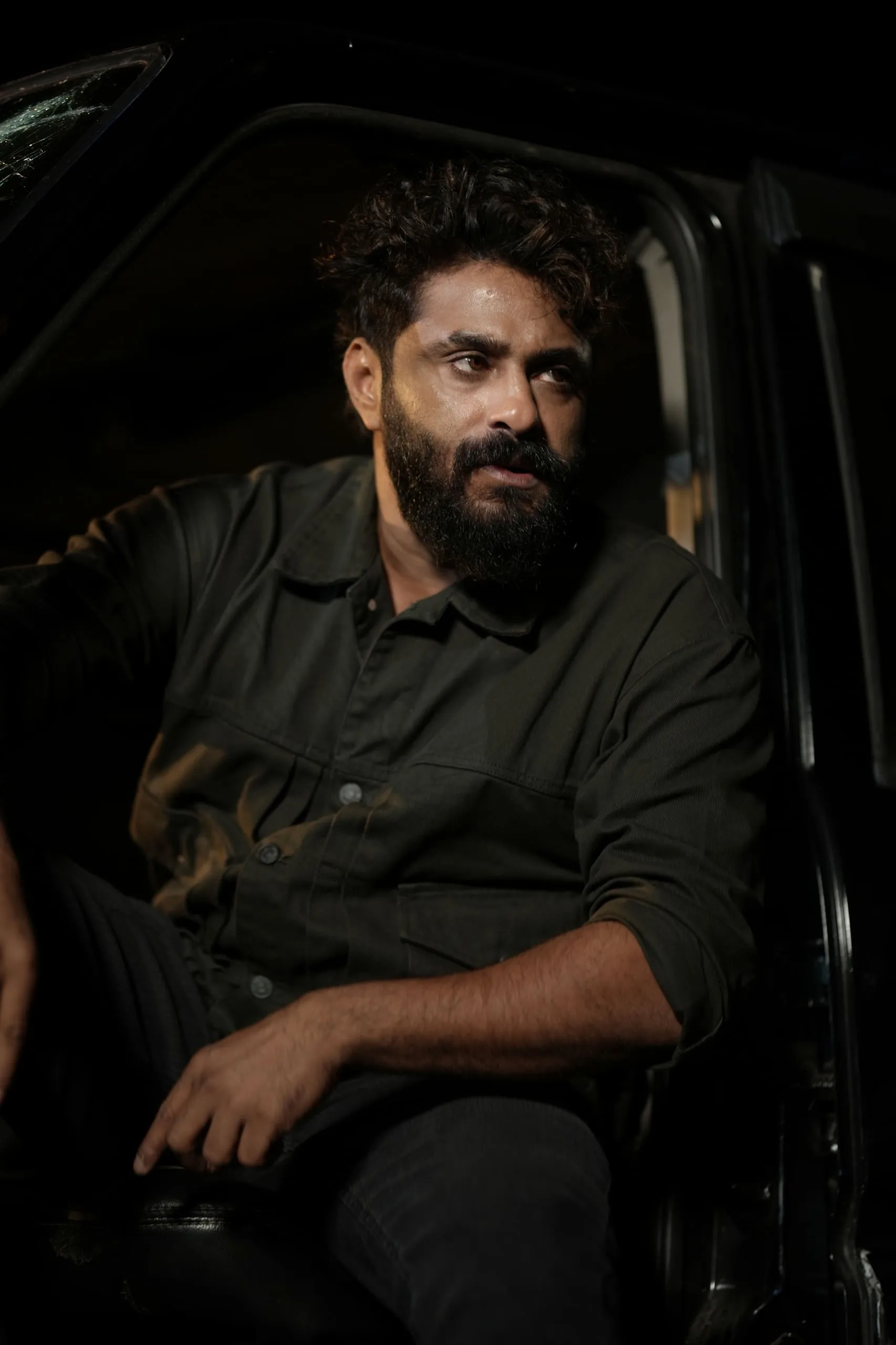 Antony Varghese in a dark green jacket, brooding inside a jeep at night in Kattalan