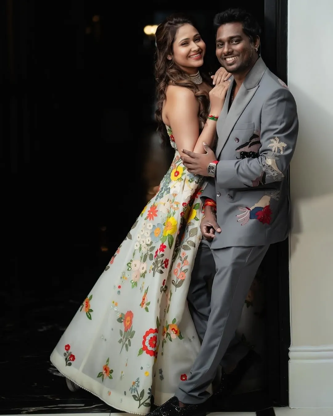 Atlee in a grey suit with painted-fabric panels and Priya in a strapless floral evening gown, posed against a dark wall for a formal portrait