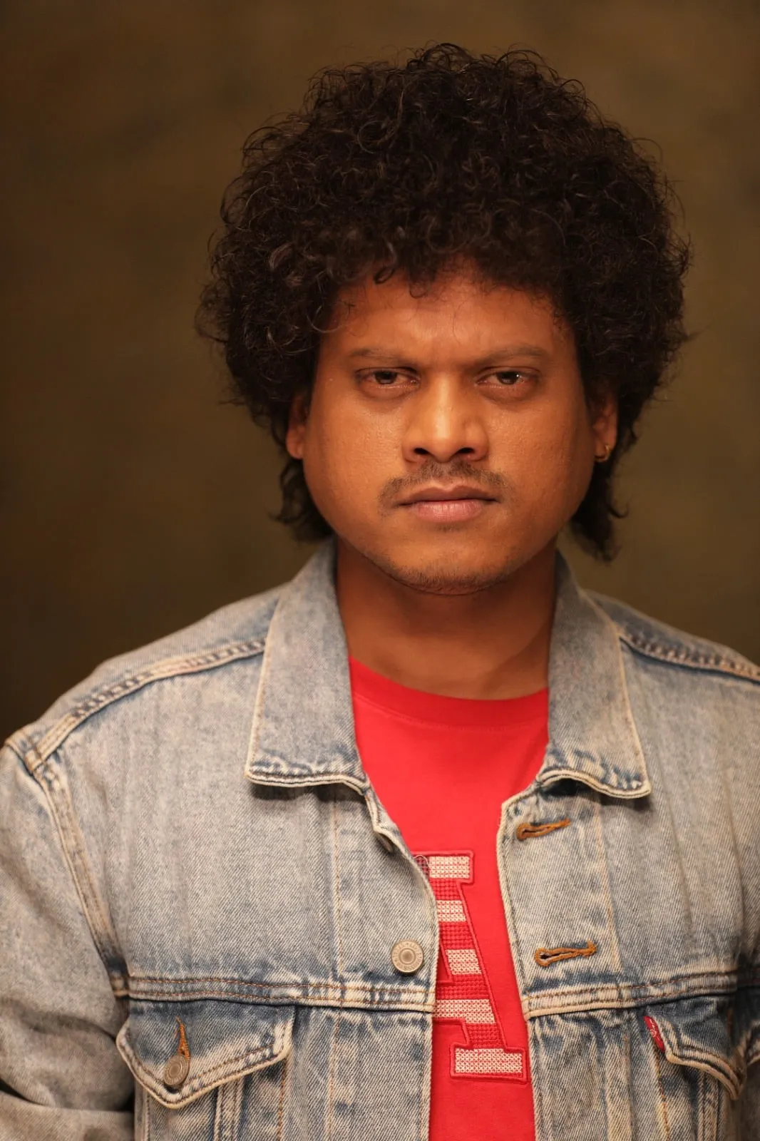 Pugazh in a denim jacket and red T-shirt, staring straight at the camera in a portrait still from 4 Idiots