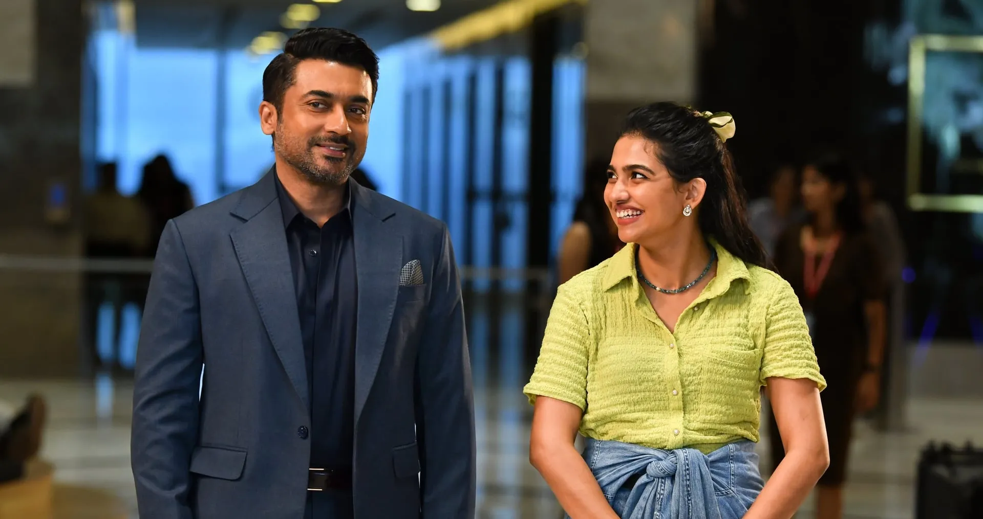 Suriya as Sanjay Vishwanath in a suit alongside Mamitha Baiju in a lime shirt inside a corporate lobby in Vishwanath & Sons