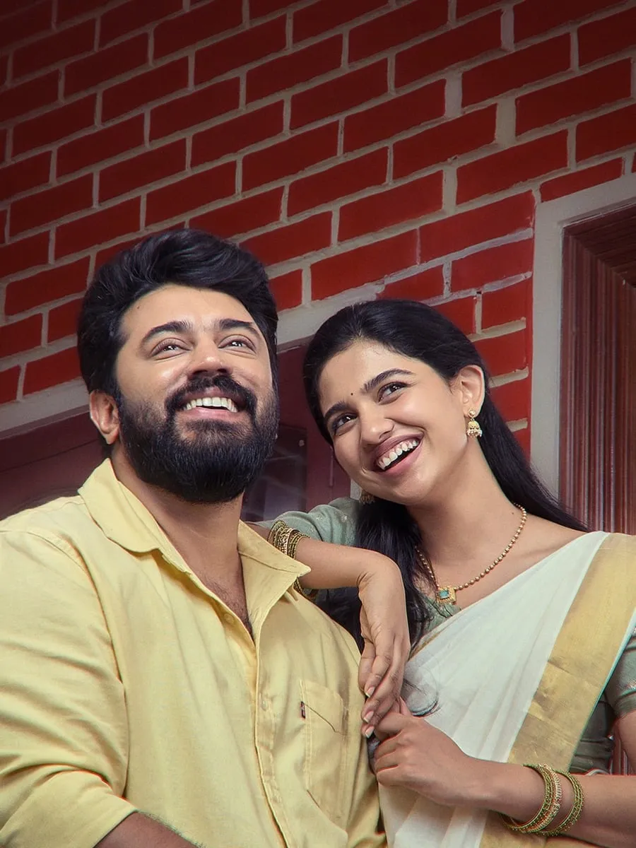 Nivin Pauly and Mamitha Baiju laugh together against a red brick wall, her arm resting on his shoulder, in Bethlehem Kudumba Unit