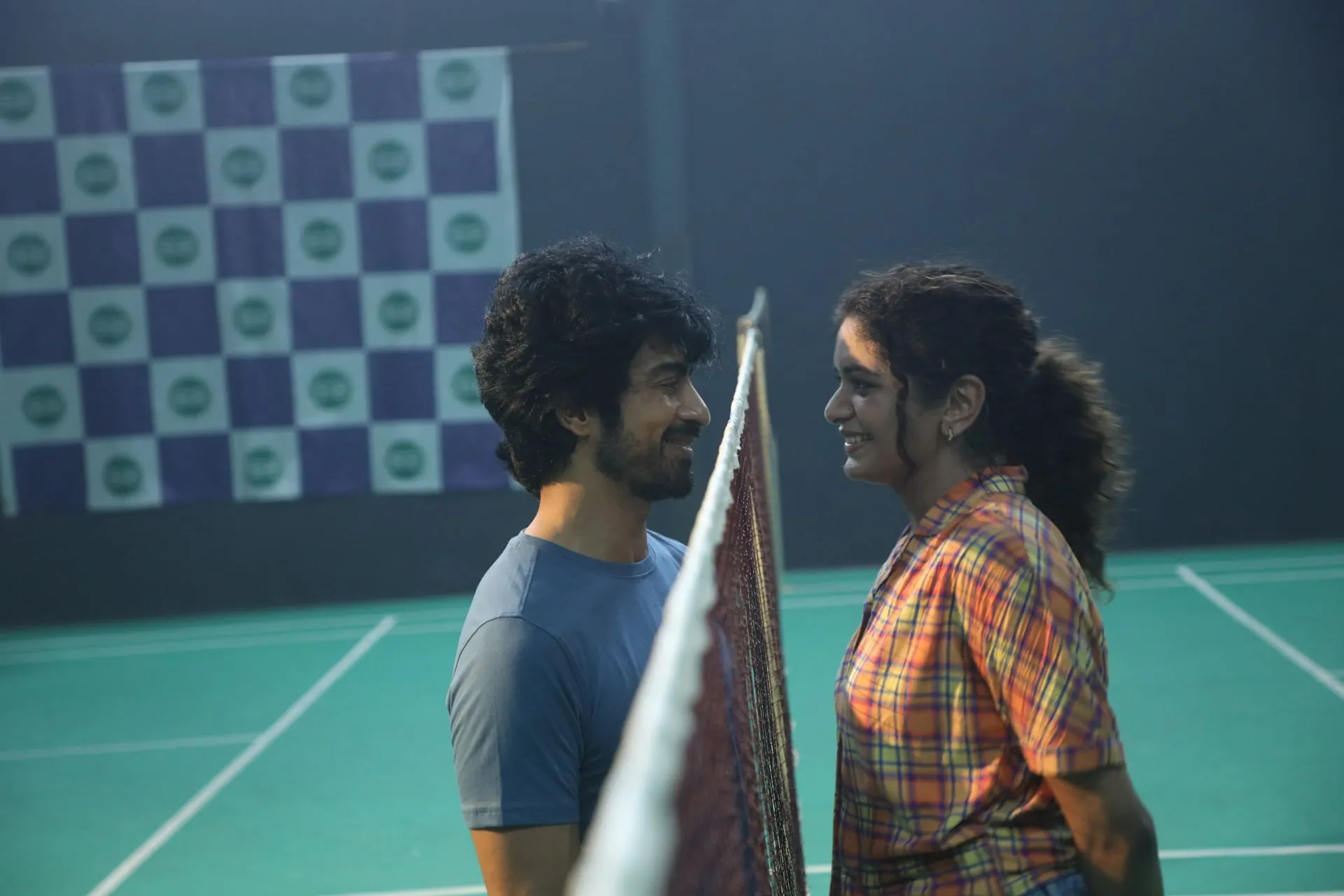 The lead pair smile at each other across the net on an indoor badminton court in Once More