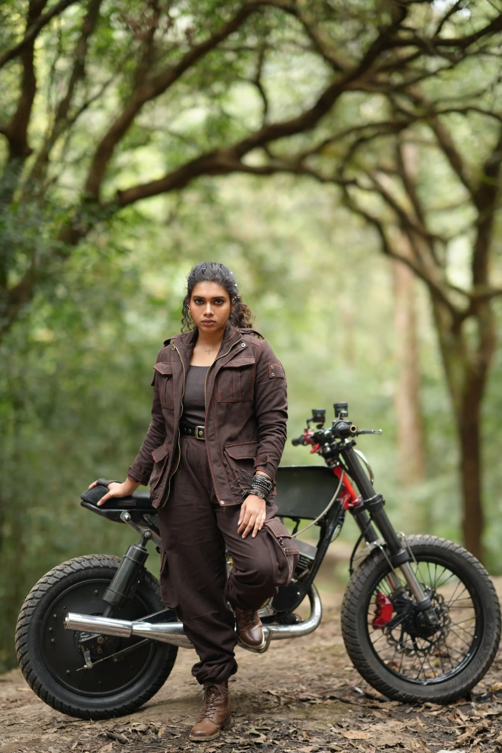 Dushara Vijayan in a brown jumpsuit standing beside a custom motorcycle on a forest trail in Kattalan