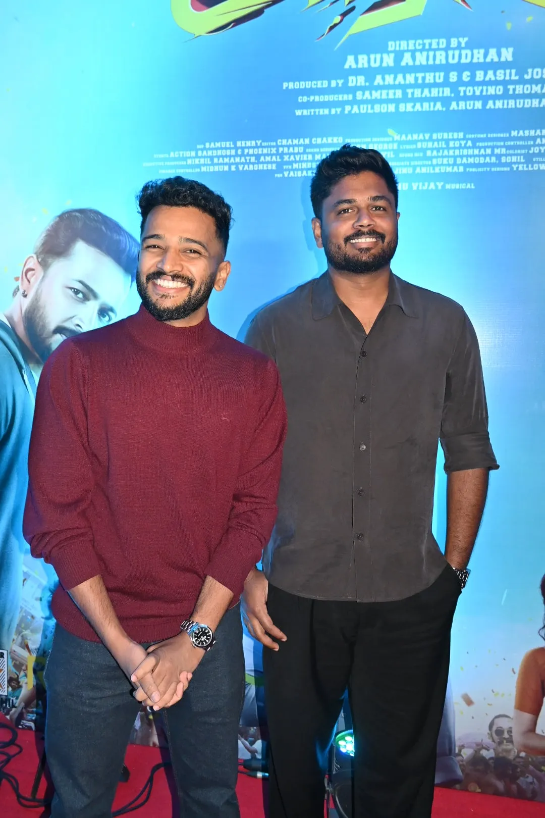 Basil Joseph in a maroon sweater and Sanju Samson in a grey shirt stand together for a photograph in front of the Athiradi credits poster at the Chennai pre-release event, both laughing