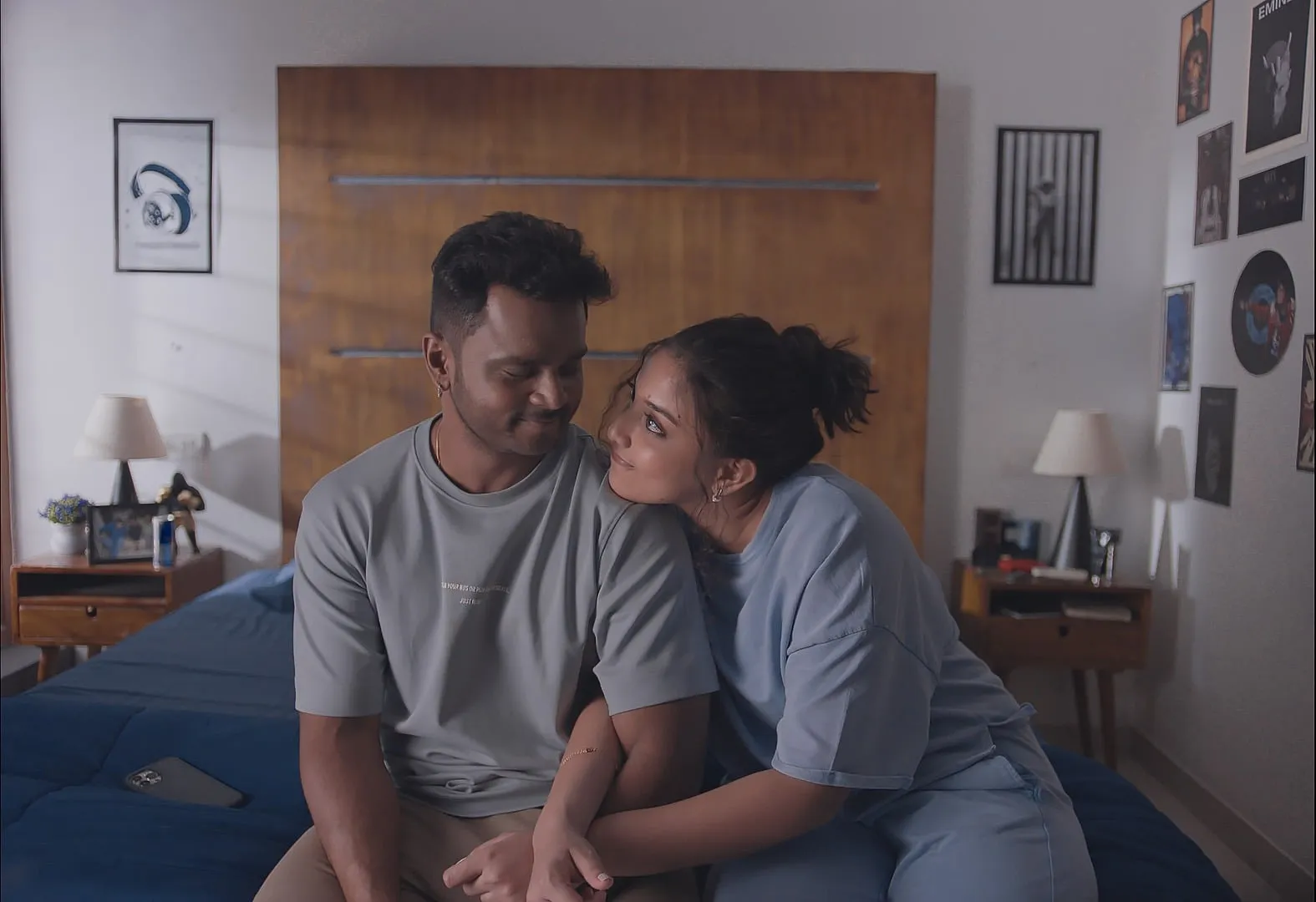 A couple sitting close on a bed in a poster-covered room in a still from Yen Ennai Yetho Seithai
