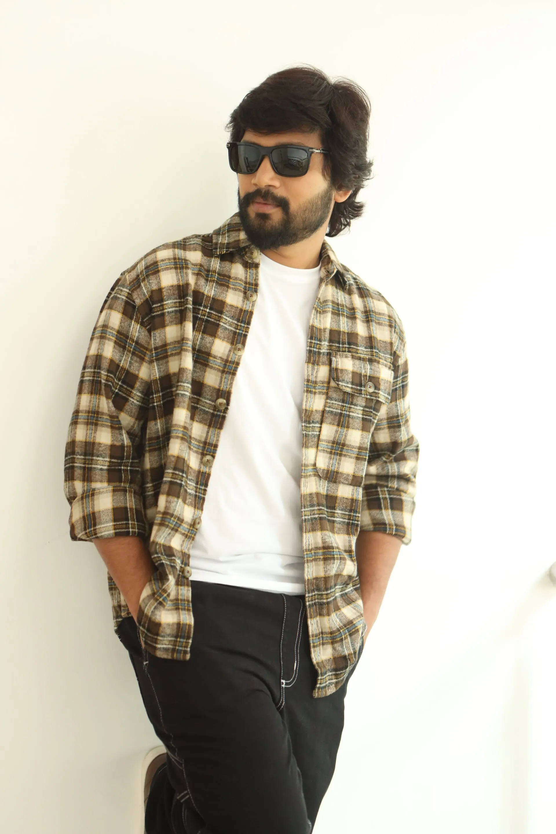 Vijay Vishwa in a plaid overshirt and sunglasses posing against a white wall