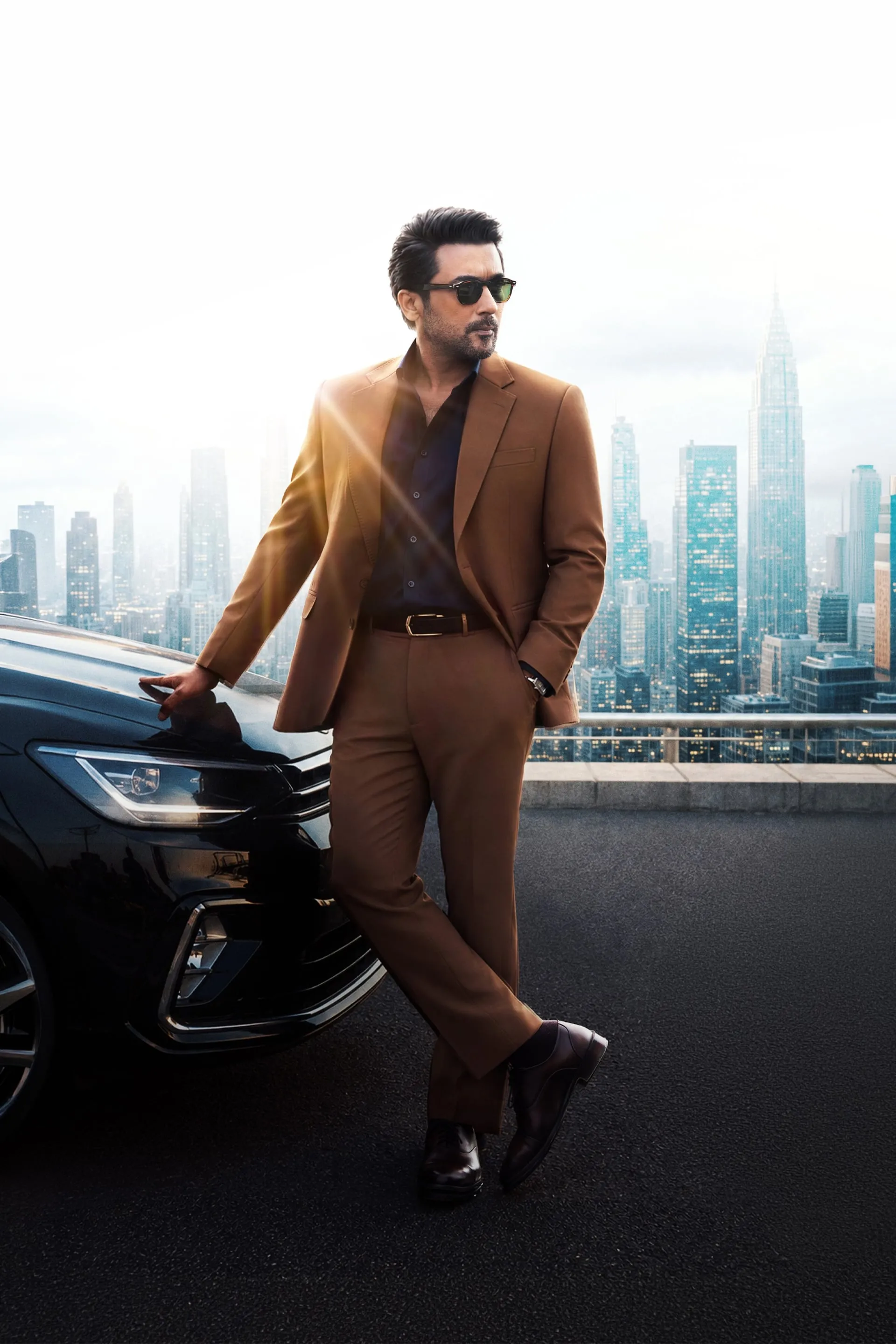 Suriya in a brown suit and sunglasses leaning against a car with a city skyline behind him