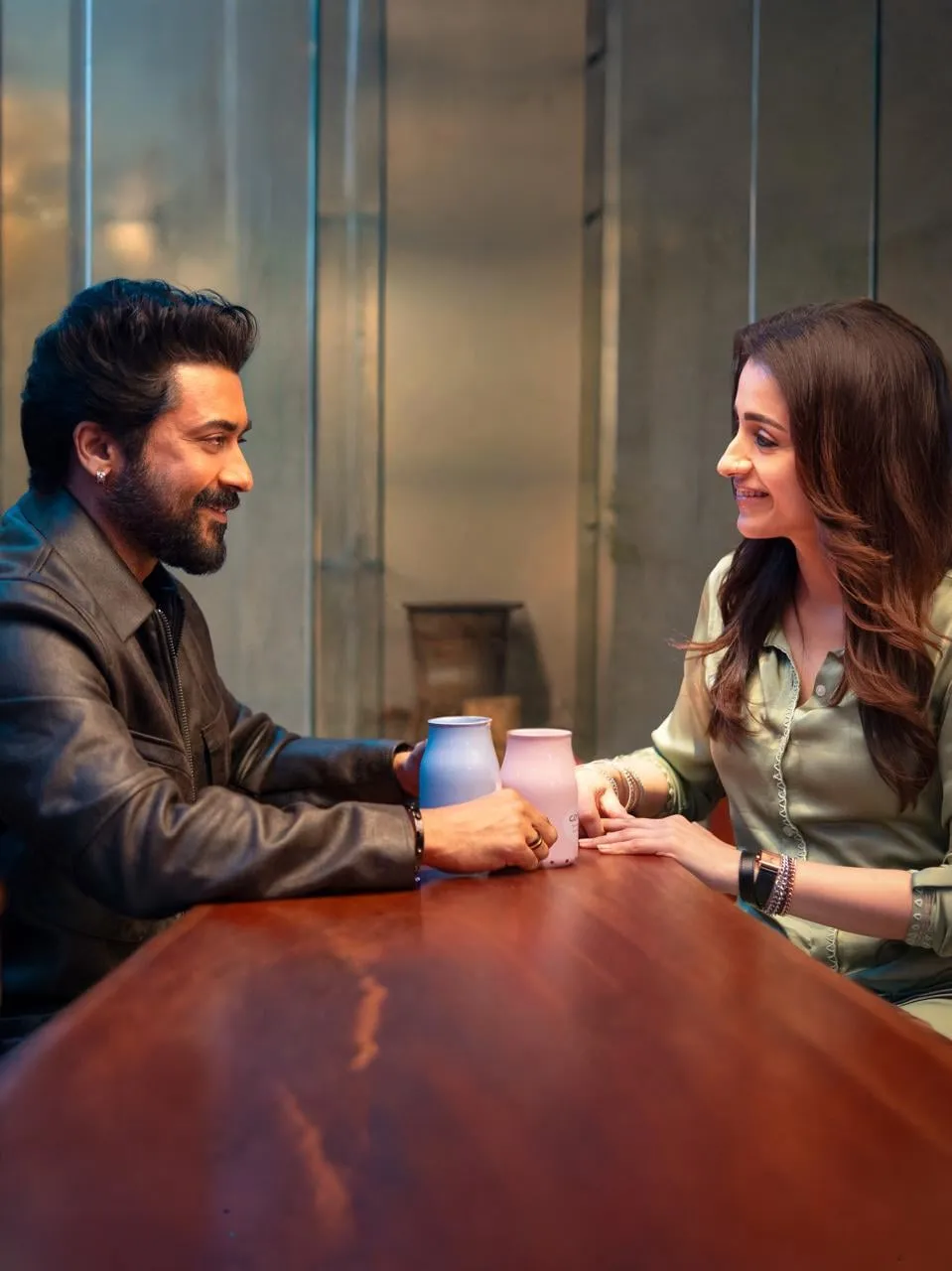 Suriya and Trisha across a wooden table in a cafe scene, both smiling at each other in a quieter beat from Karuppu