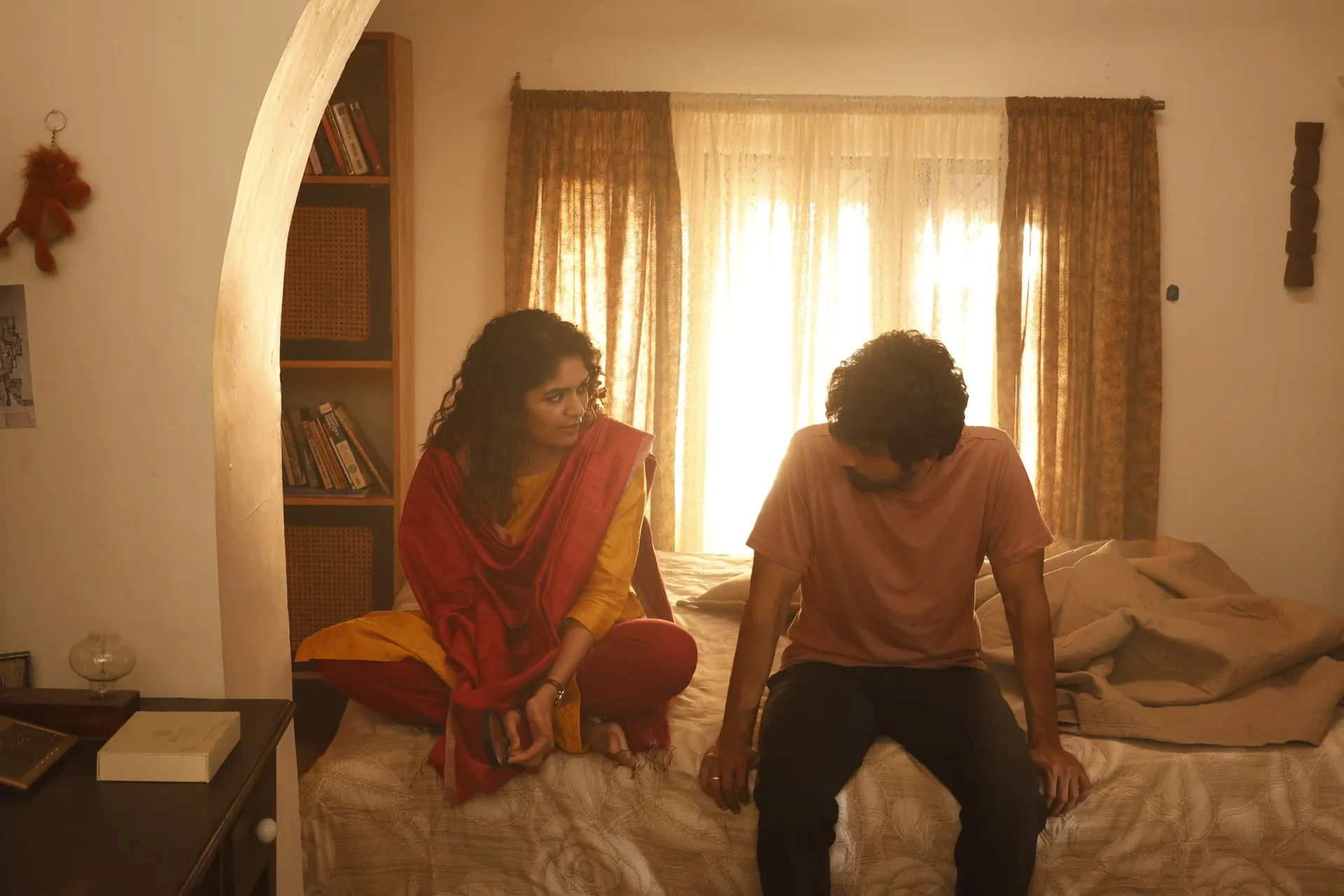 Anjali, in a yellow kurta, sits on a bed trying to read a dejected Raghu, who stares at the floor in Once More