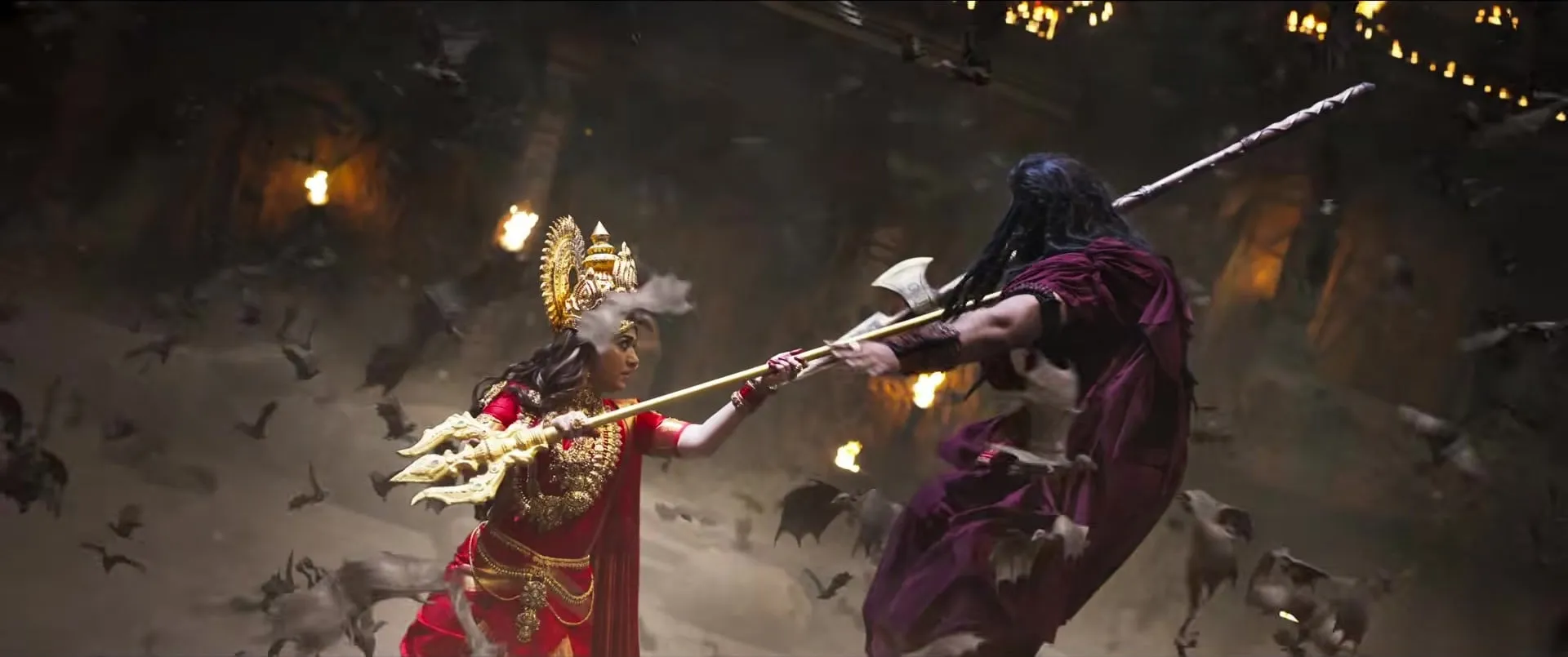 The goddess, trident raised, clashes with a dark-robed figure in a cave lit by torches