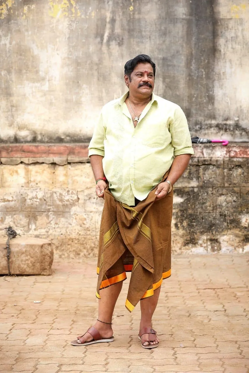 Vettai Muthukumar standing in a lime shirt and checked lungi against a weathered wall