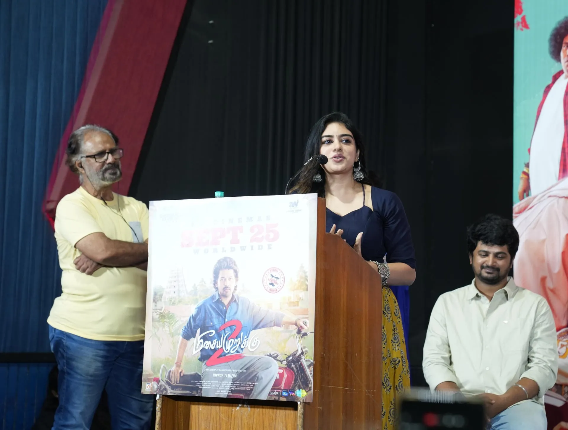 A speaker in a blue top and yellow patterned skirt at the Meesaya Murukku 2 microphone